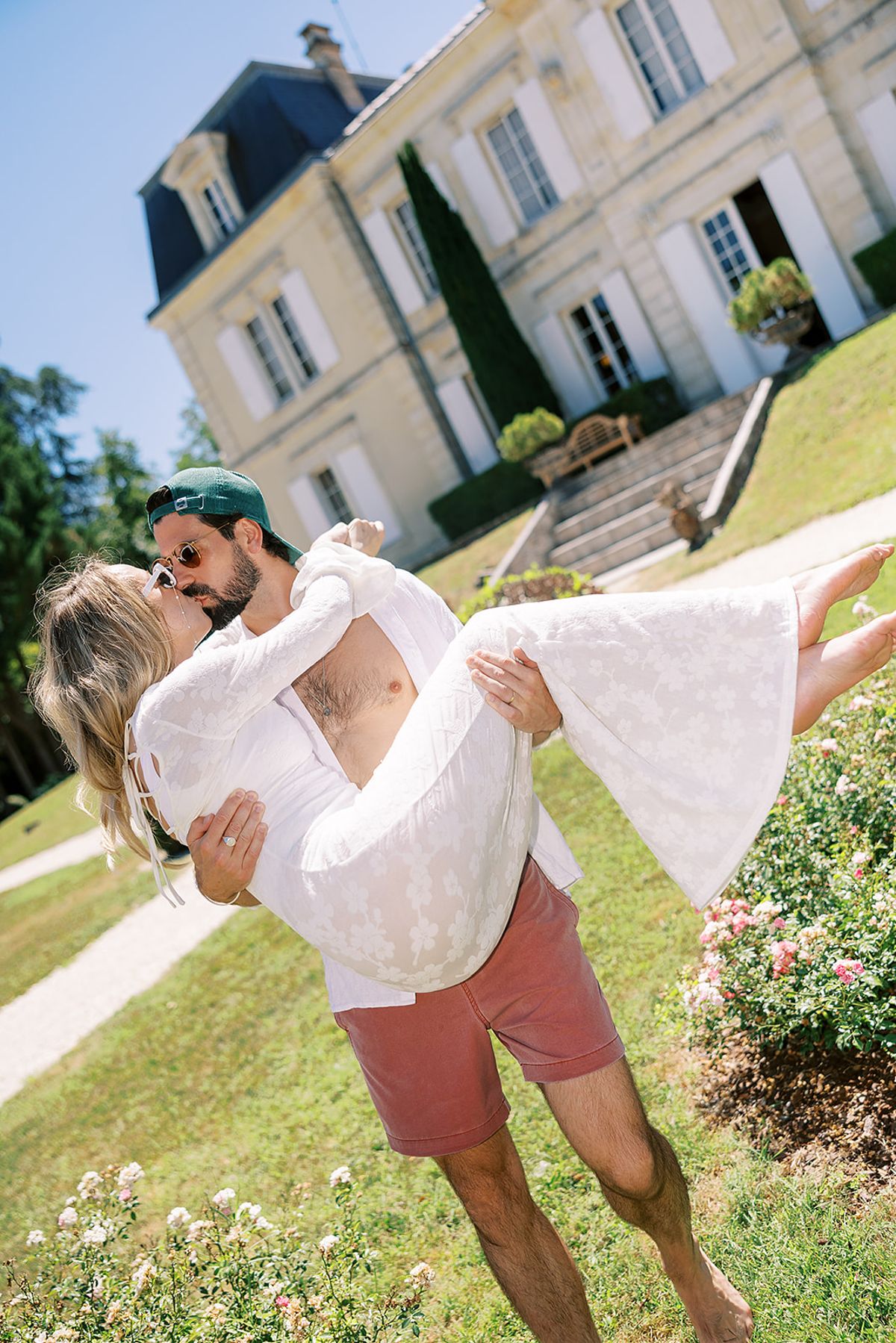 The groom, in shorts and a cap, carries the bride in his arms on a lawn in front of a stone chateau.