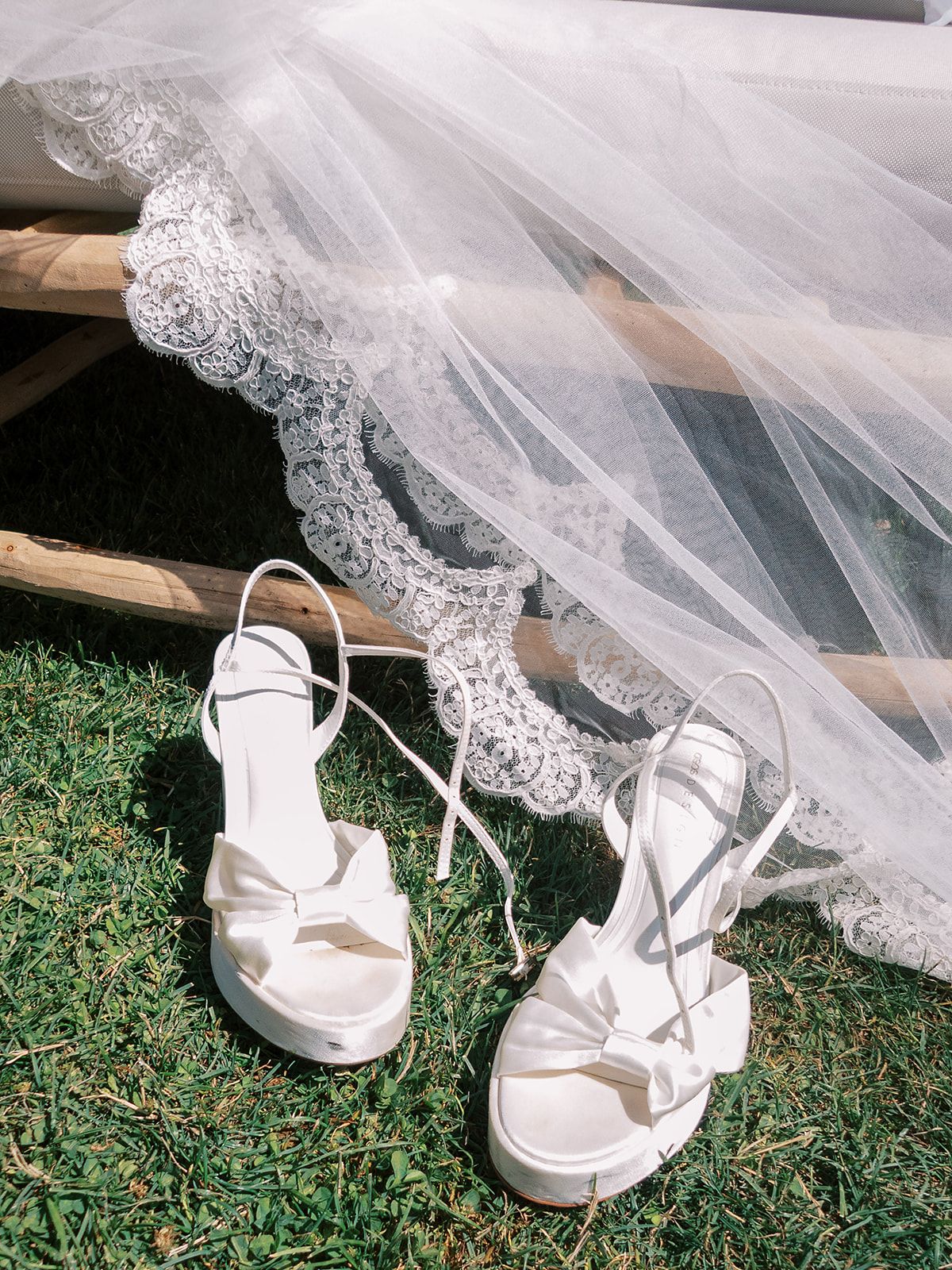 A pair of white satin platform sandals with bows and ankle ties on grass, beside a lace-edged bridal veil.