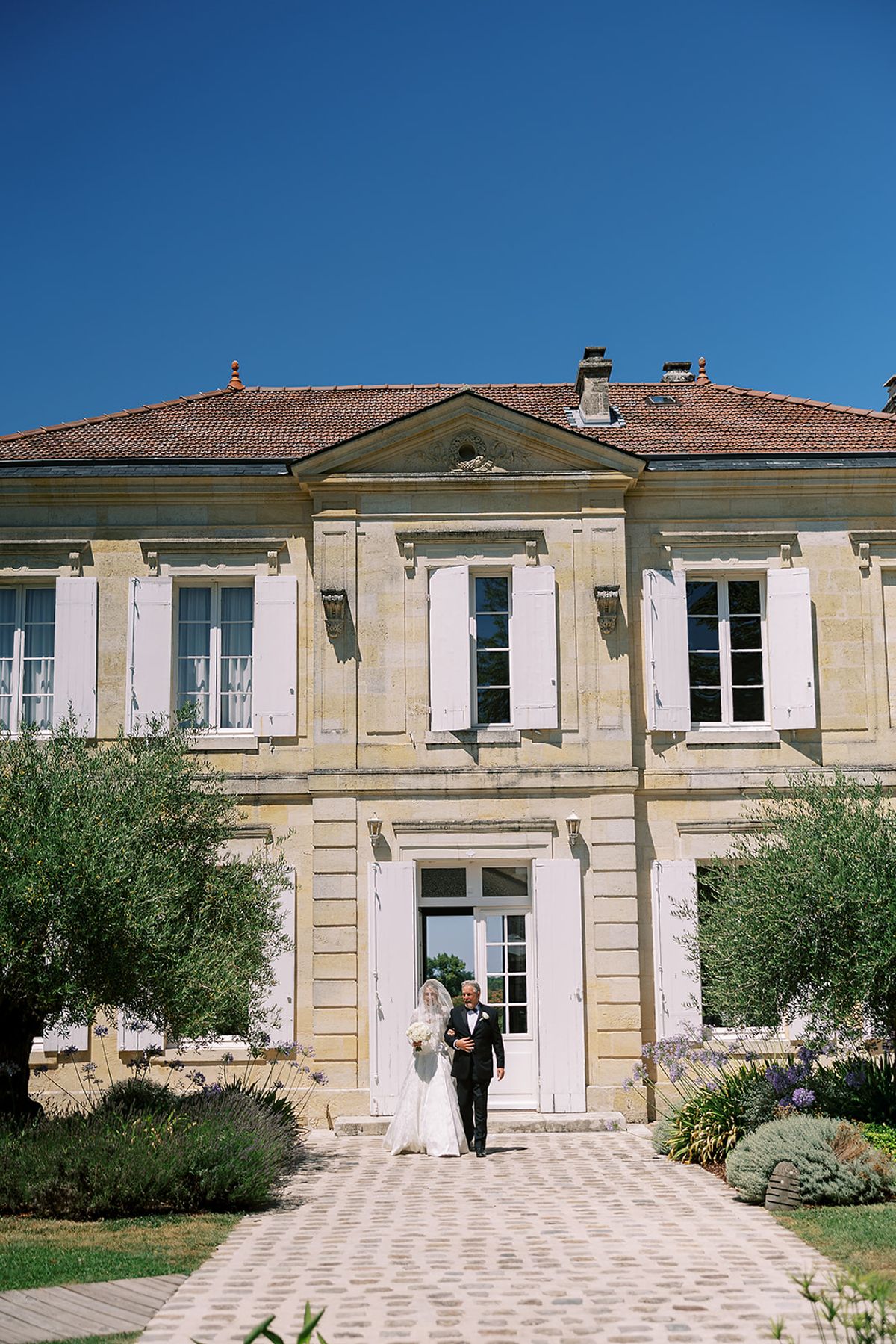 A bride in a white gown and veil walks arm in arm with a man in a dark suit from a stone manor house along a cobbled path.