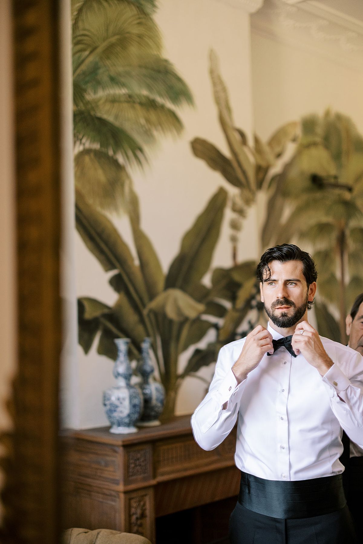 The groom, in a white shirt and black cummerbund, adjusts his bow tie before a palm-leaf mural and blue-and-white vases.