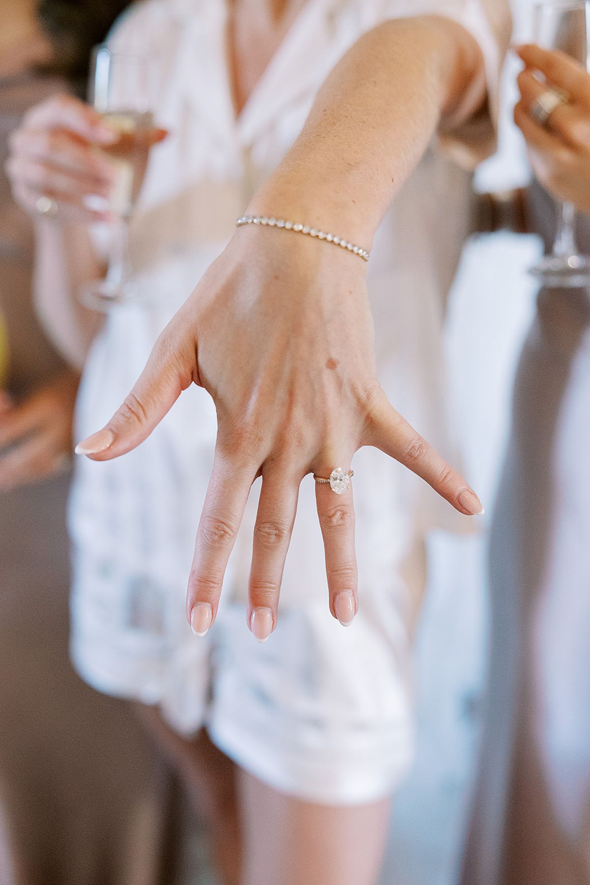 A woman in white pyjamas holds out her hand to show an oval ring, as guests with champagne glasses stand blurred behind.