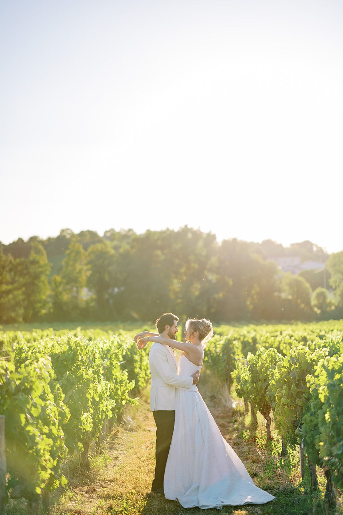 The bride in a long white gown and the groom embrace between rows of vines, with bright sunlight glowing behind them.