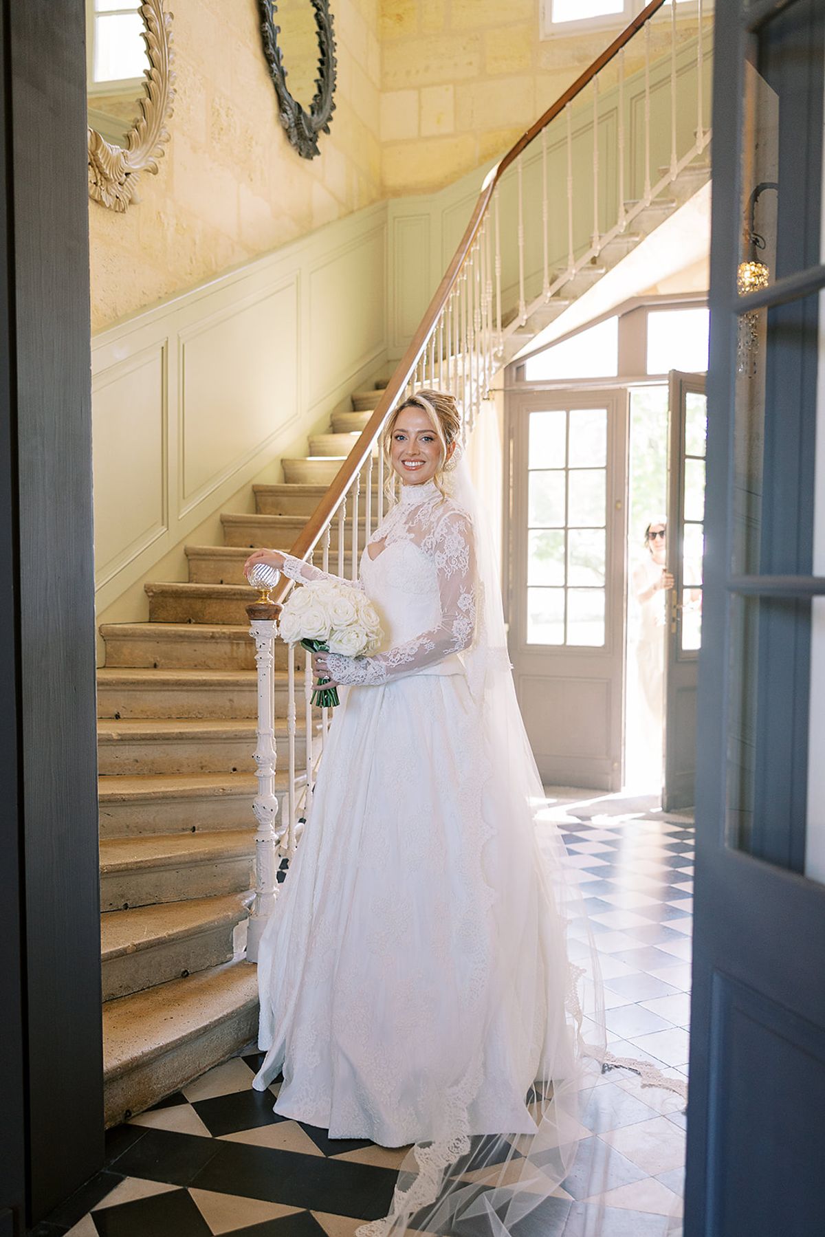 The bride in a long-sleeved lace gown holds a white bouquet in a doorway beside a wooden staircase and chequered floor.