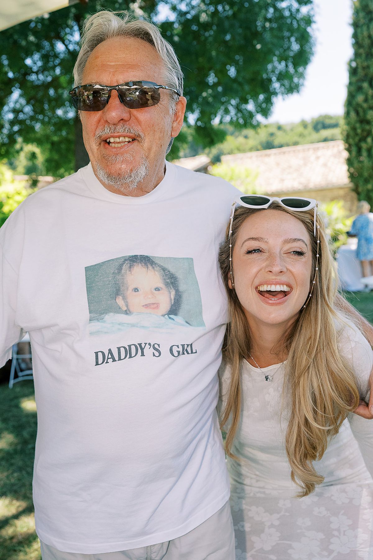 A man in sunglasses wearing a white T-shirt with a printed baby photograph stands beside a laughing woman in white, in a garden.