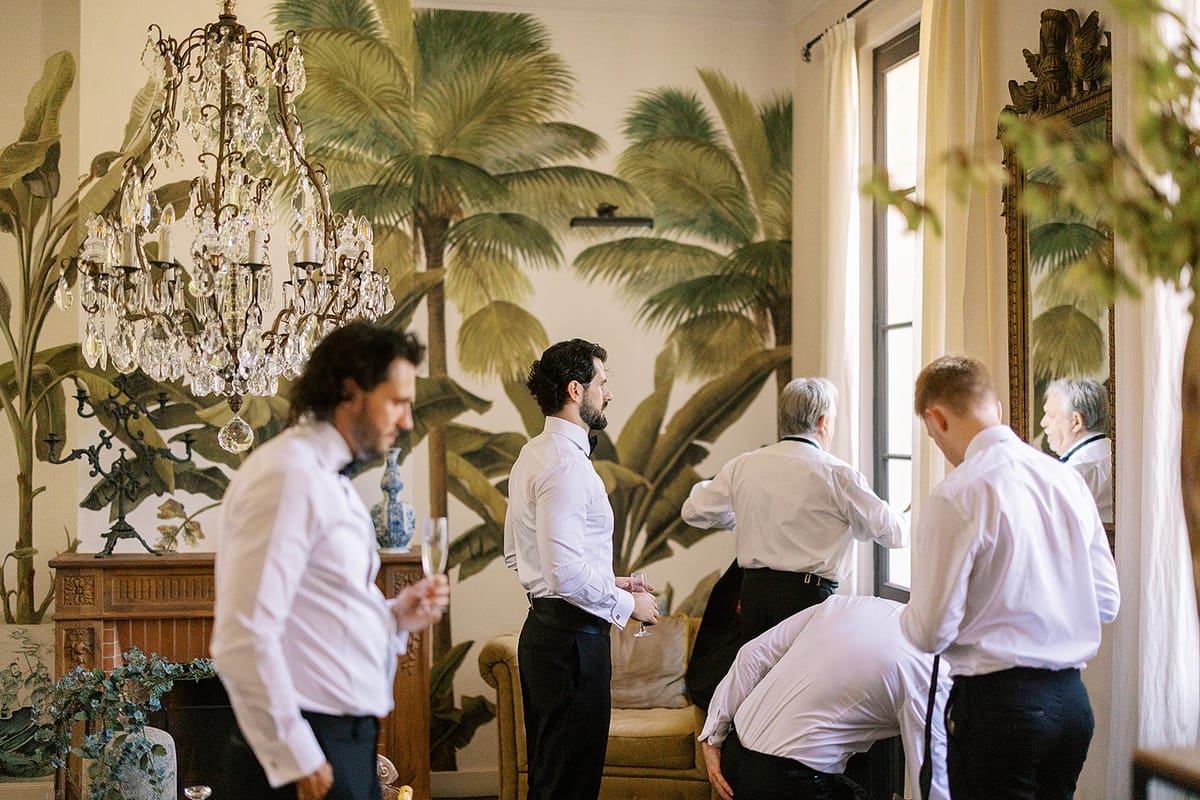 Several men in white shirts stand and bend in a room with palm-patterned wallpaper and a crystal chandelier, getting dressed together.