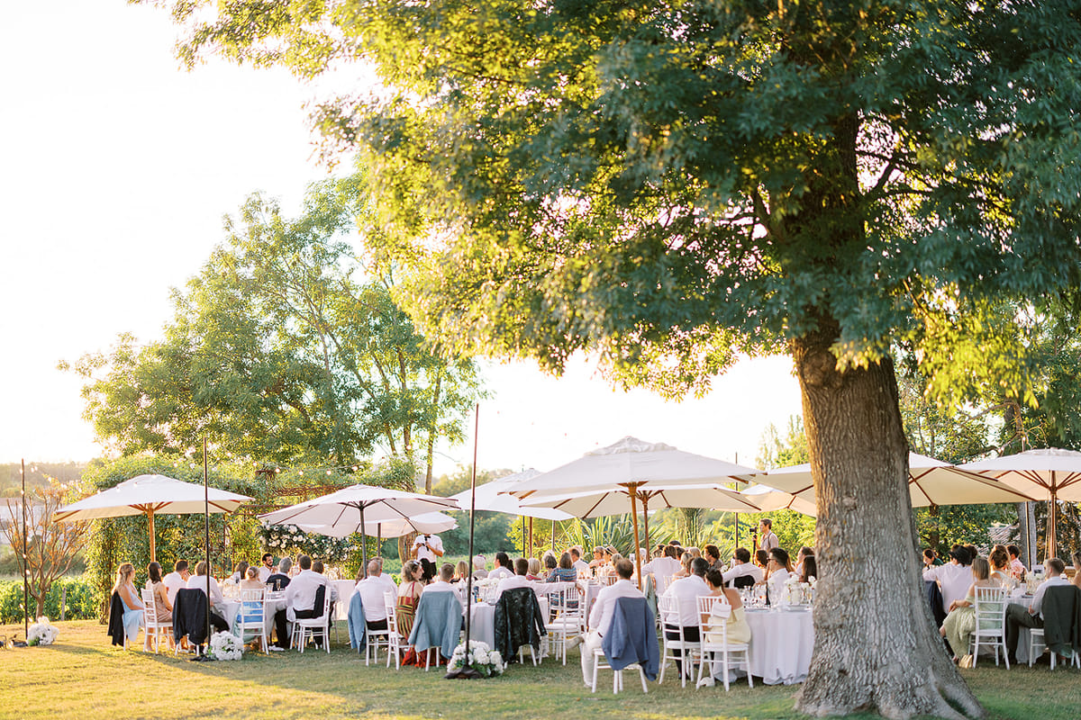 Guests sit at long tables beneath white parasols on a lawn, shaded by a large tree in warm, low sunlight.