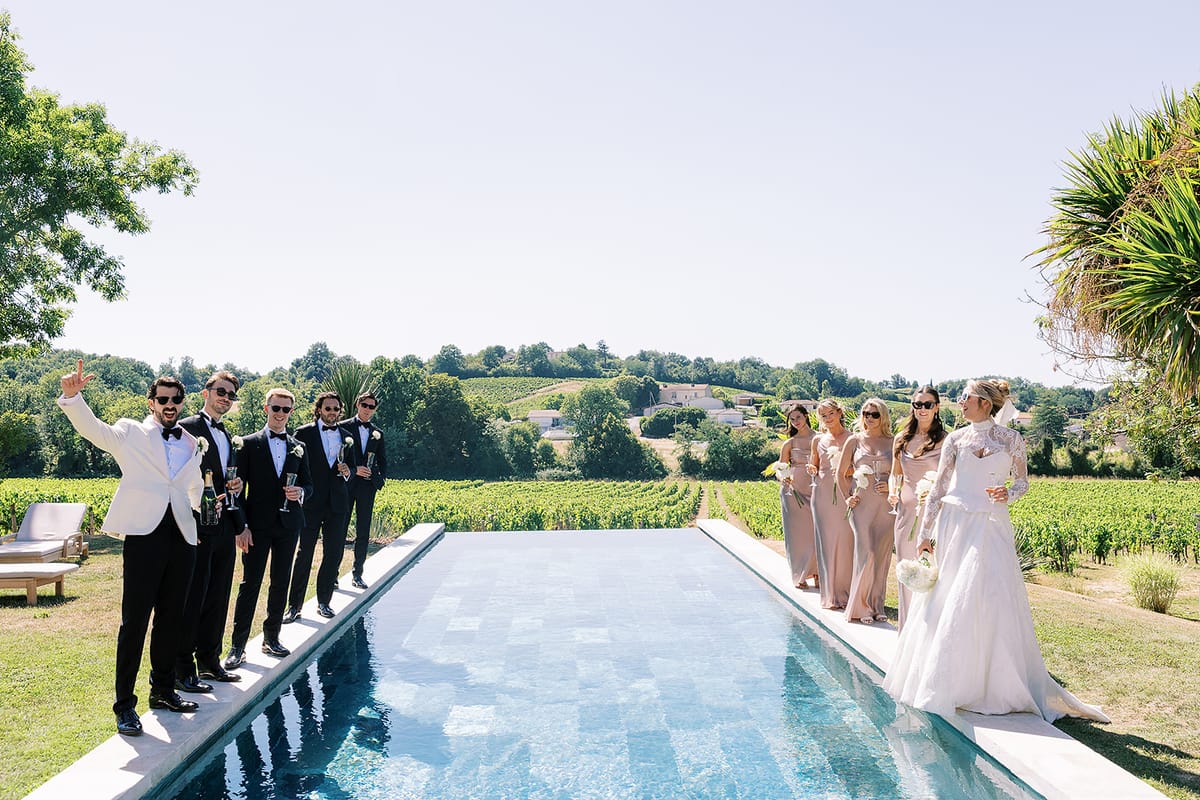 Groomsmen and bridesmaids stand in two lines along the edge of a long pool, with the bride at the far end.