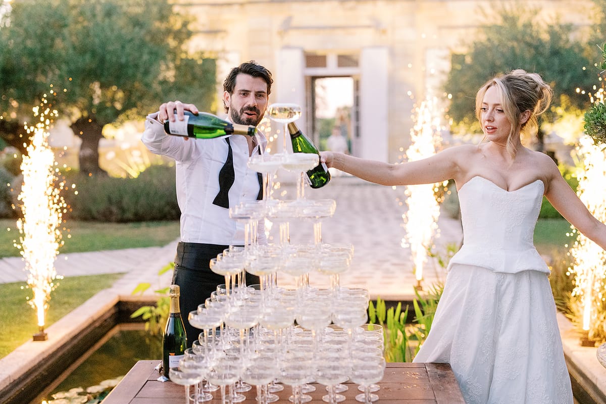The bride in a white strapless dress and the groom in a white shirt pour champagne over a glass tower, sparklers behind.