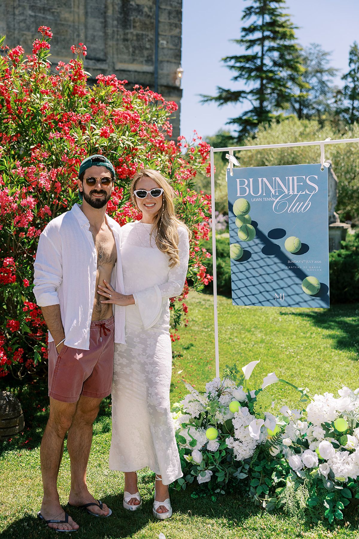 The couple stand arm in arm on a lawn in front of red roses, white flowers and a blue sign reading Bunnies Club.