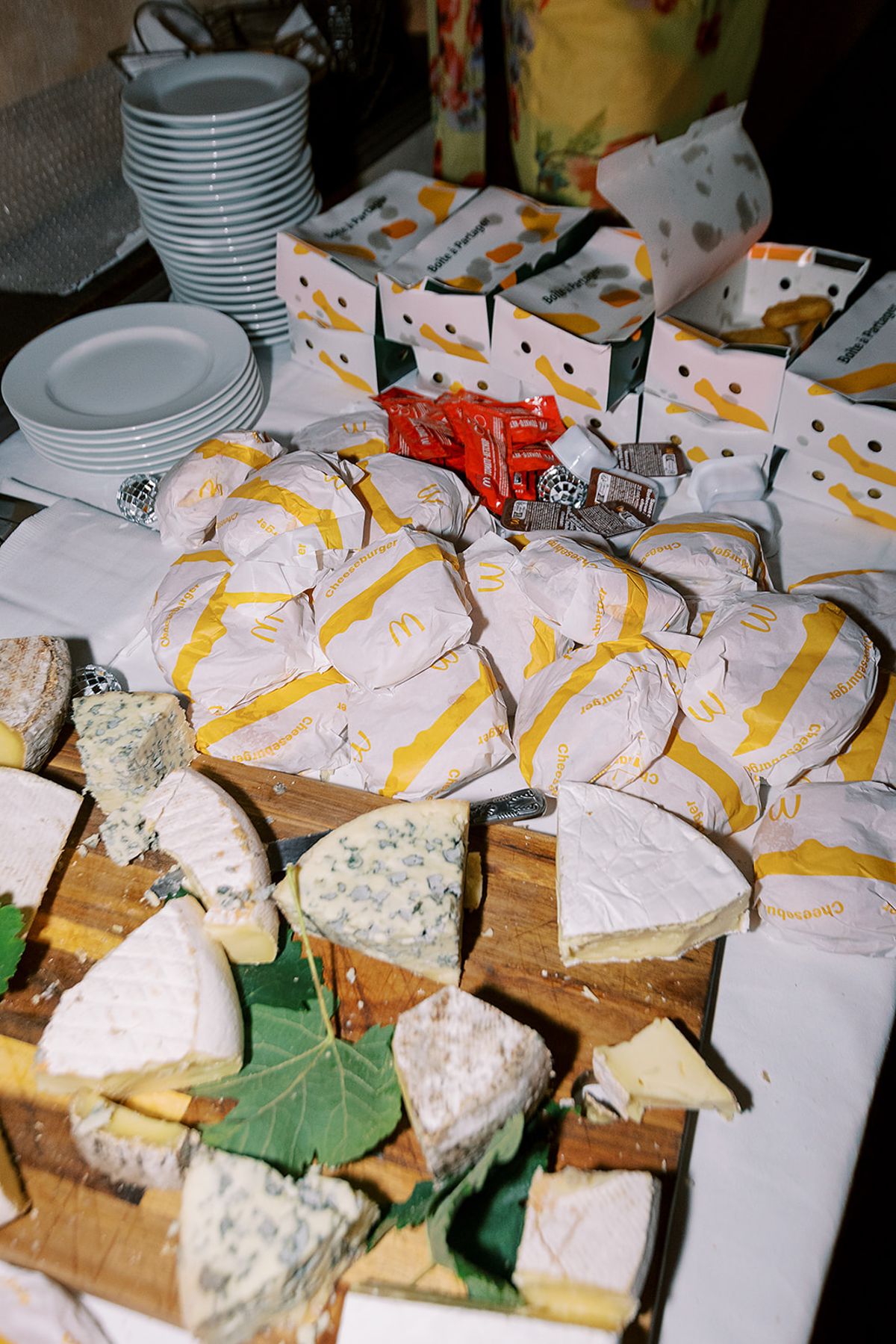 A table holds stacked plates, boxes, cheeses wrapped in yellow-patterned paper and cut wedges of blue cheese on a wooden board.