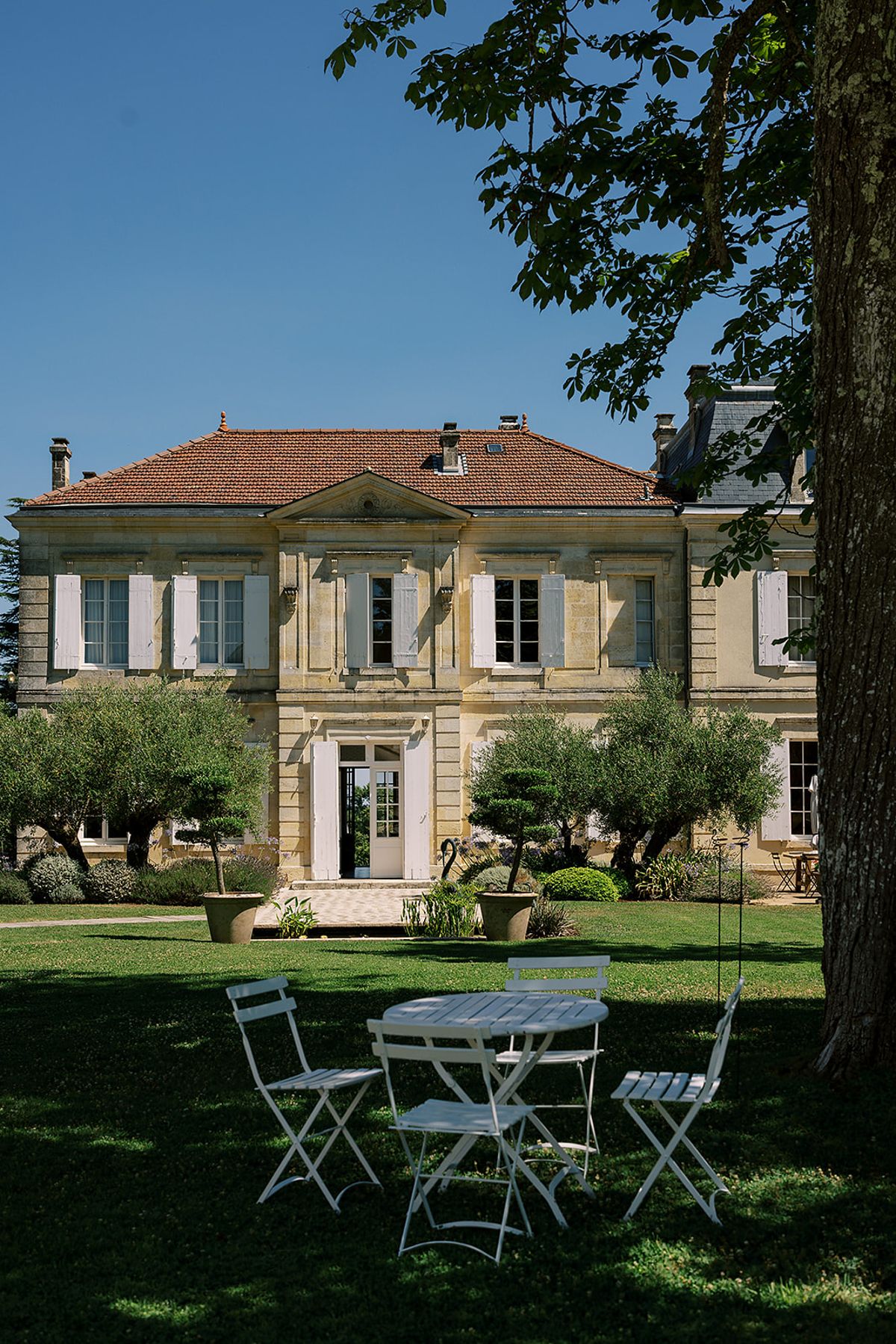 A stone chateau with a terracotta roof and shuttered windows stands behind a lawn with olive trees and a small table with white folding chairs.