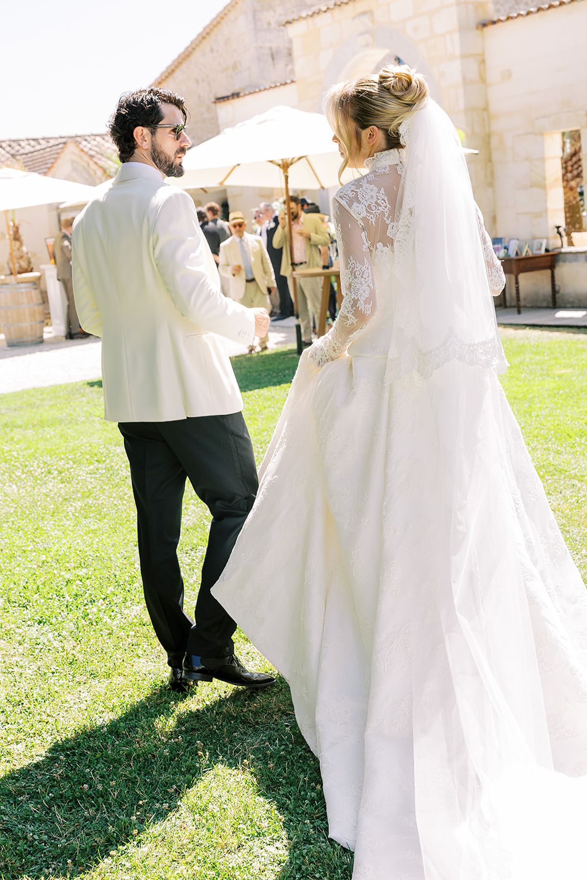 The bride in a long veil and lace gown stands beside the groom in a white jacket on a sunlit lawn.