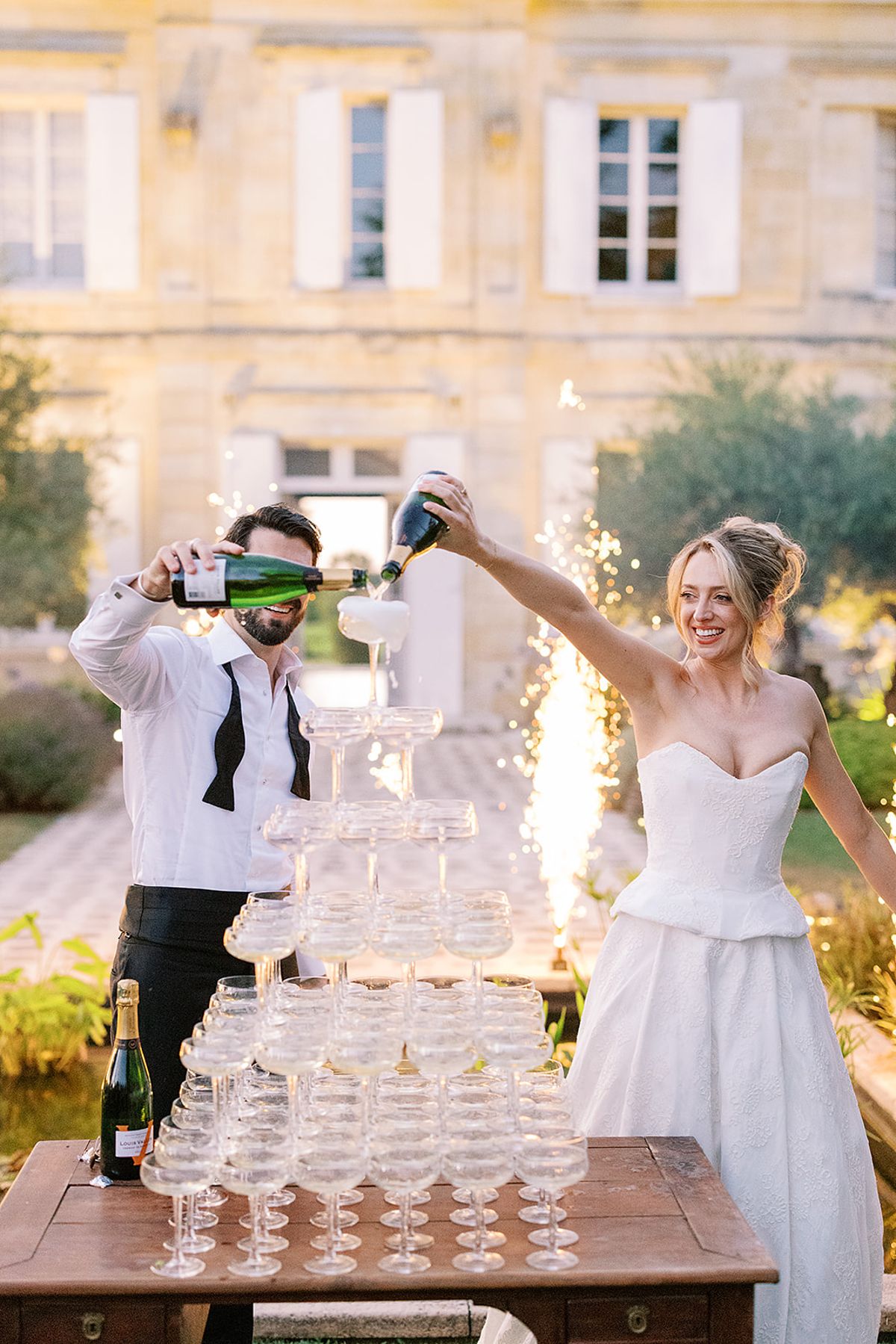 The bride and groom pour champagne from two bottles onto a tower of coupe glasses, with sparklers and a stone château behind.