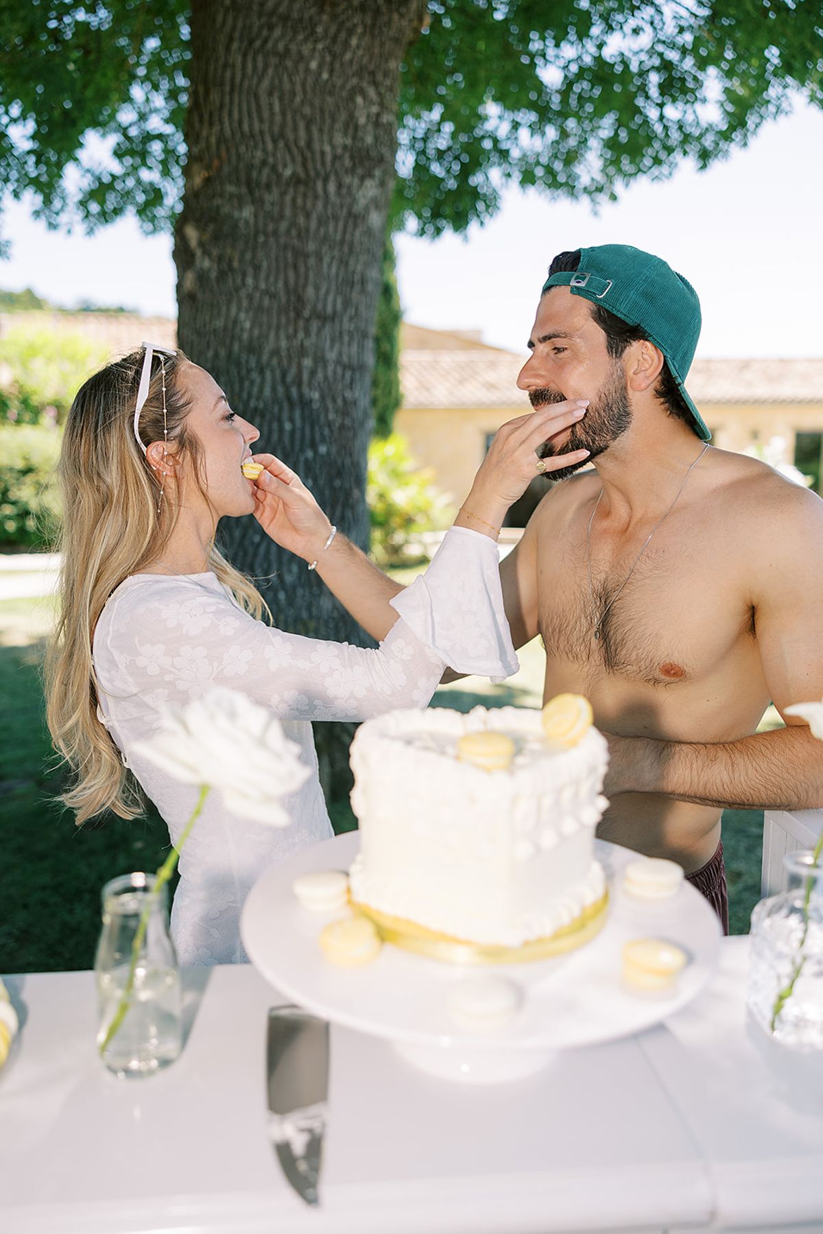 A couple feed each other macarons beside a heart-shaped white cake on a table under a tree.
