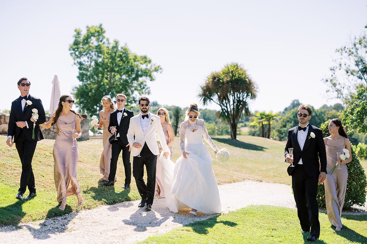 The couple walk along a sunny path on a lawn, surrounded by smiling guests in suits and pastel dresses.