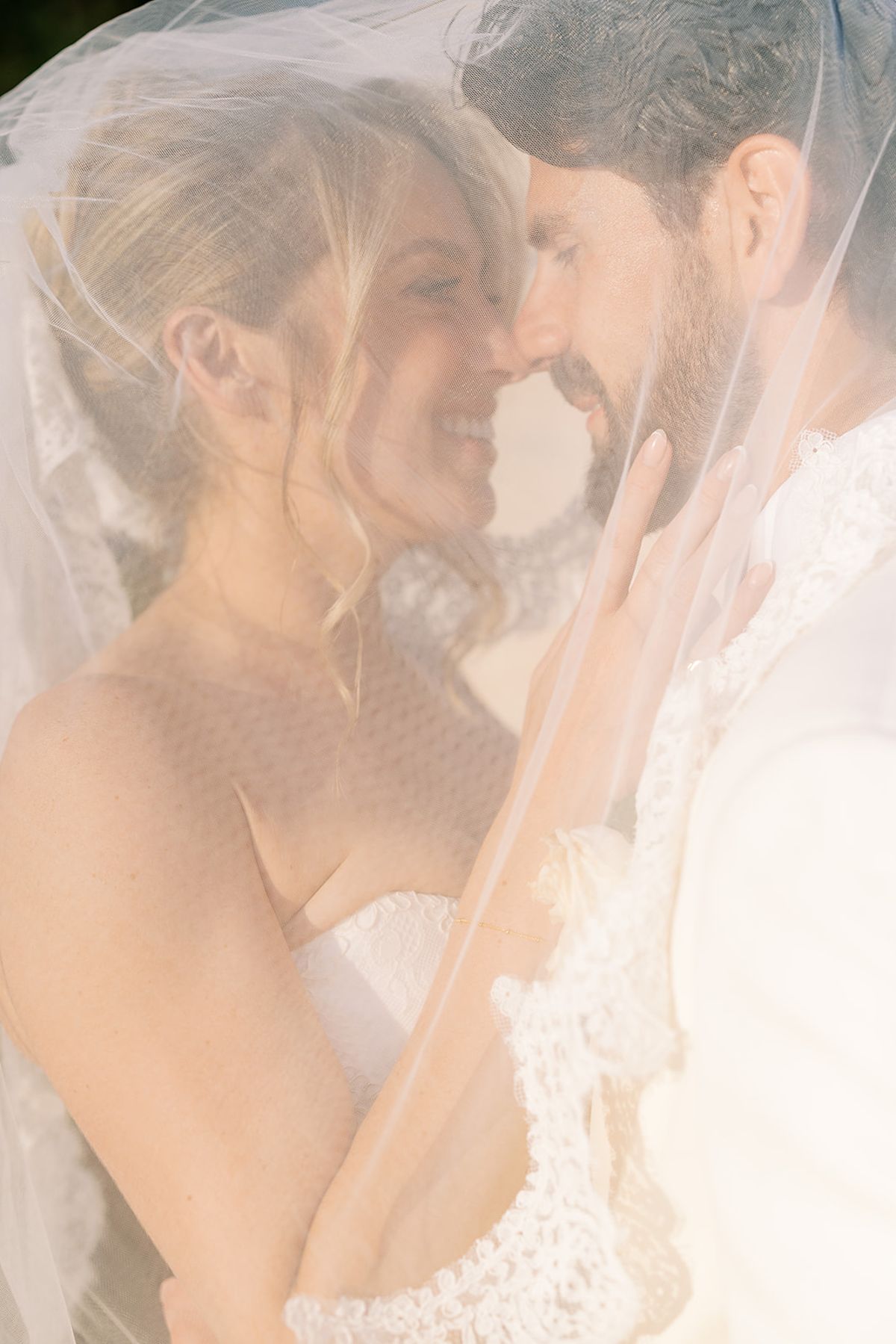 The bride and groom stand forehead to forehead beneath a sheer veil in soft, glowing light.