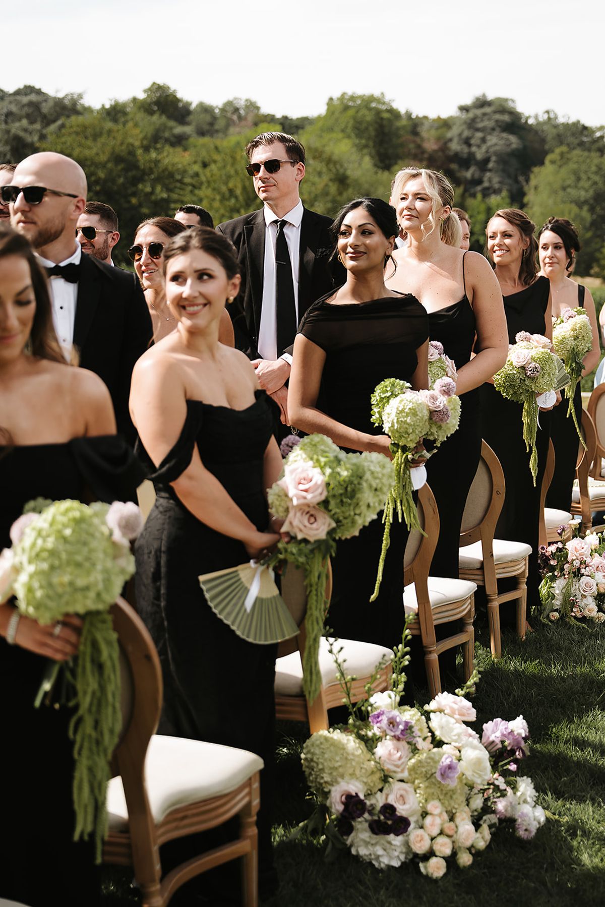 Bridesmaids in black dresses hold green and white bouquets as guests stand beside rows of wooden chairs on the lawn.