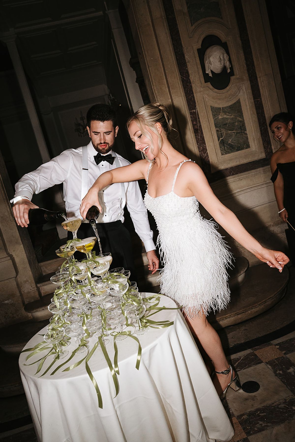 A man in a bow tie and a woman in a short beaded feather dress pour champagne into a coupe tower tied with green ribbons, in a marble hall at night.