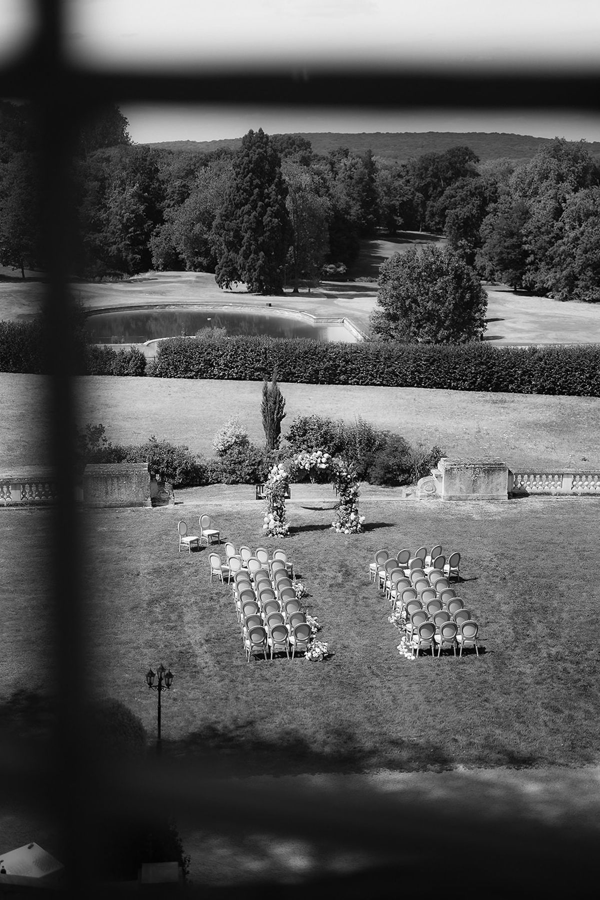 A black and white view through a window frame over parkland and a lawn where rows of chairs are arranged before a flower arch.