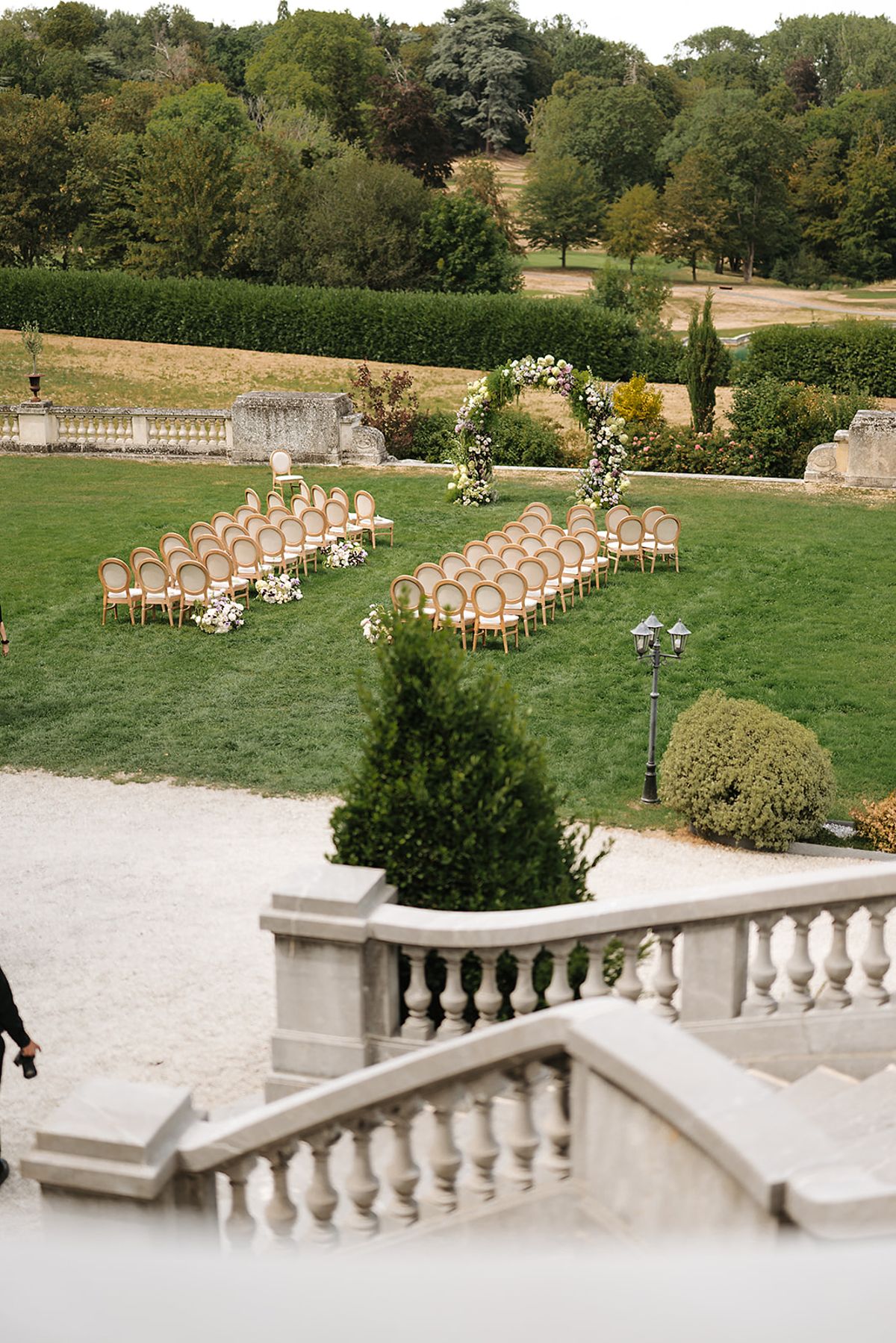 Rows of wooden chairs face a floral arch on a lawn beside a stone balustrade, with a person partly visible at the left edge.