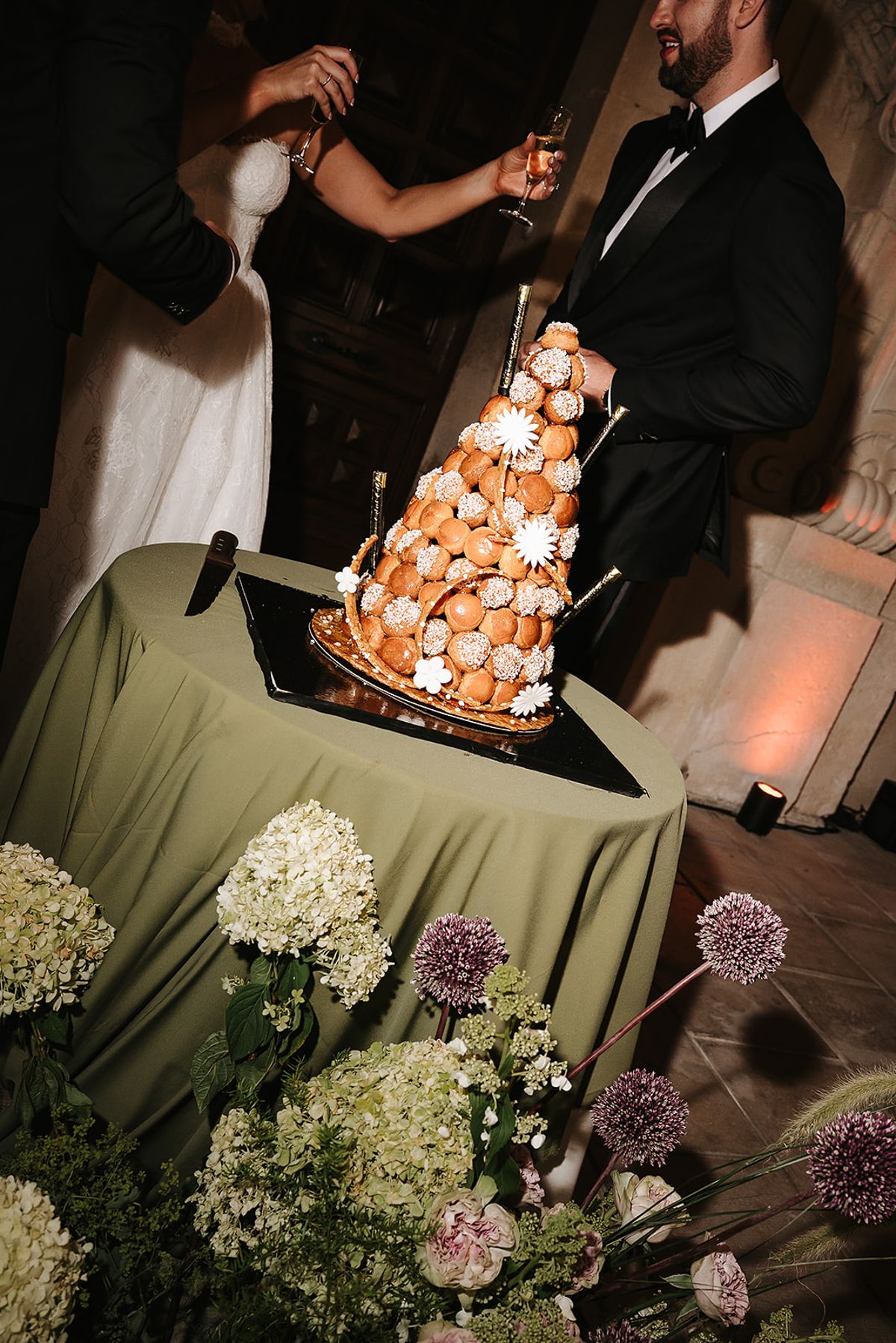 The groom and another person cut a tall tower of choux pastries on a table with a green cloth and flowers.