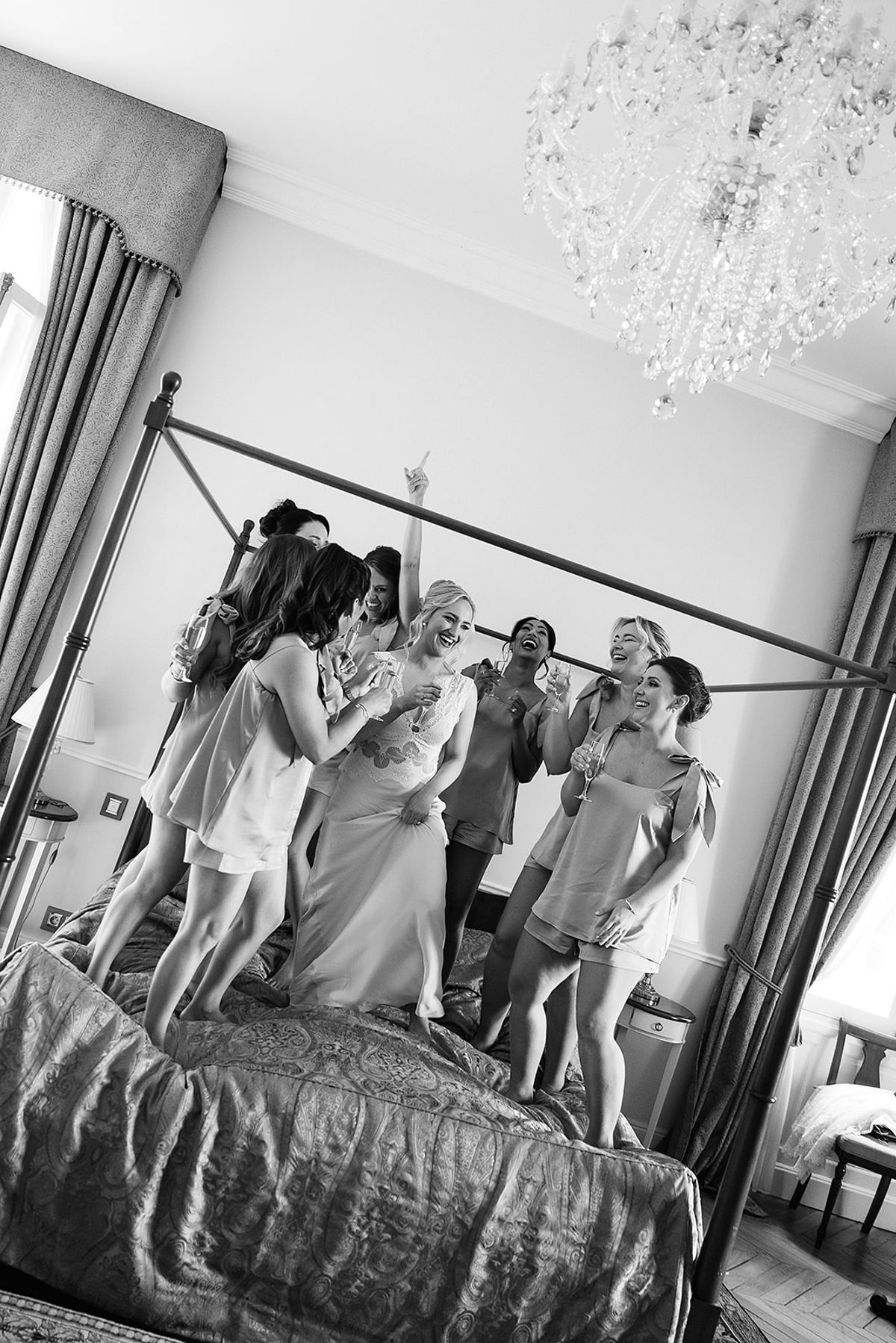 In black and white, a group of women in matching robes stand close together on a bed beneath a crystal chandelier.