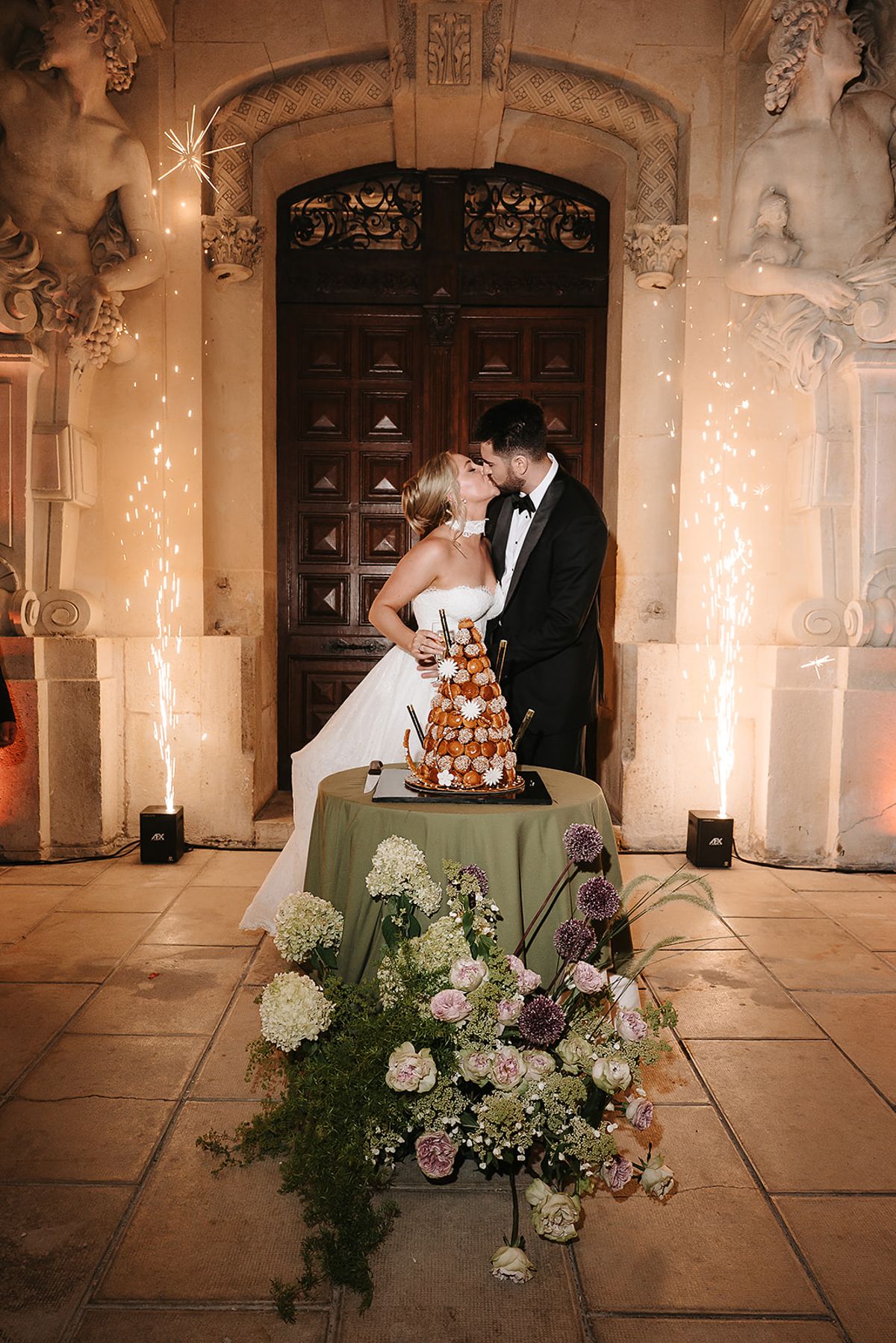 The bride and groom kiss in front of a carved wooden door beside a tall cake, with sparklers glowing at either side.