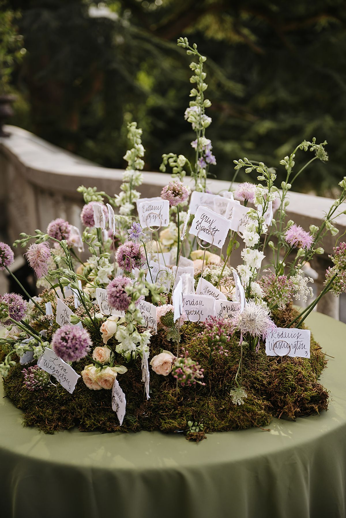A table display of lilac and white flowers set in moss, with small handwritten tags attached to the stems.