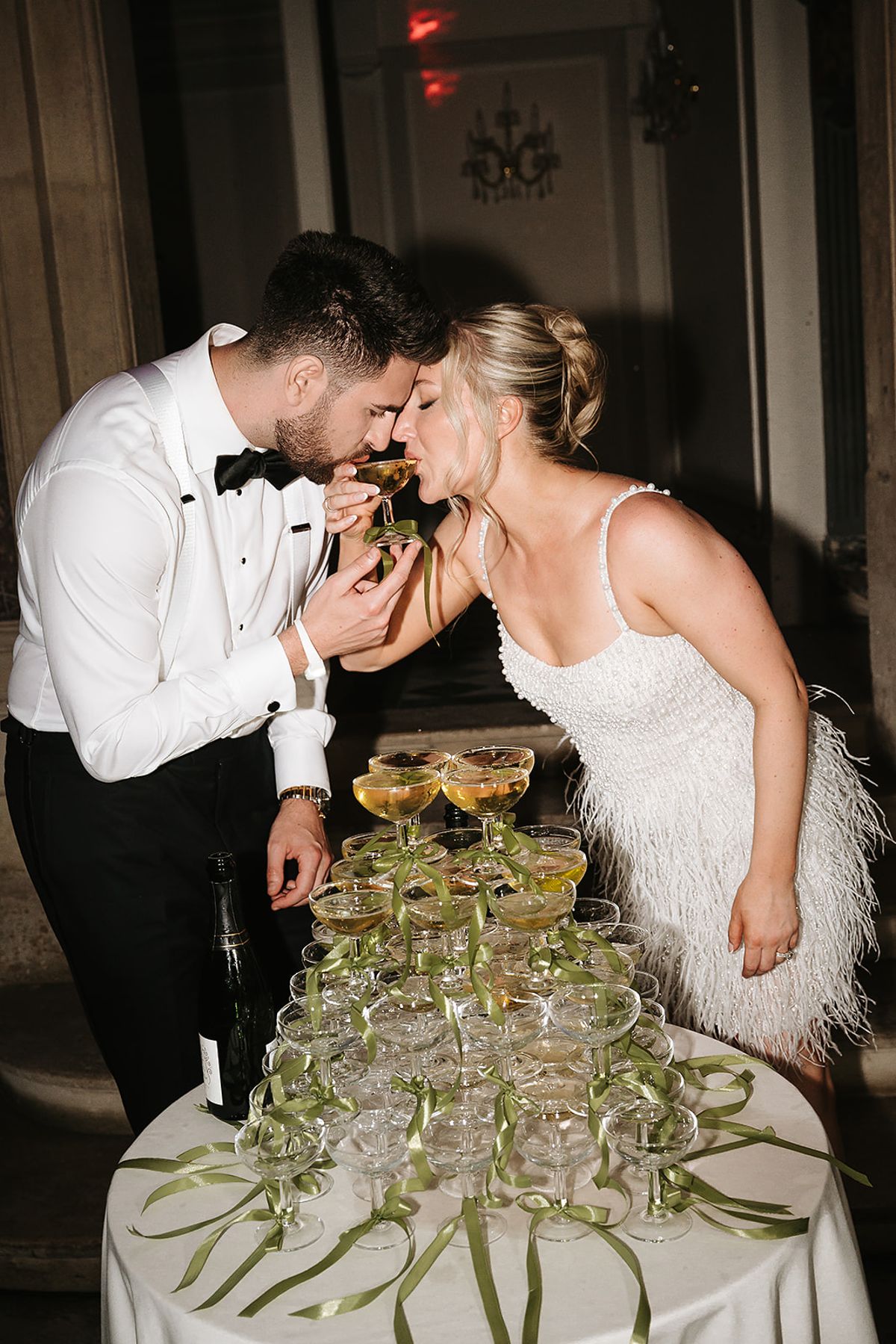 The groom in a bow tie and the bride in a feathered white dress sip from one glass above a champagne coupe tower.