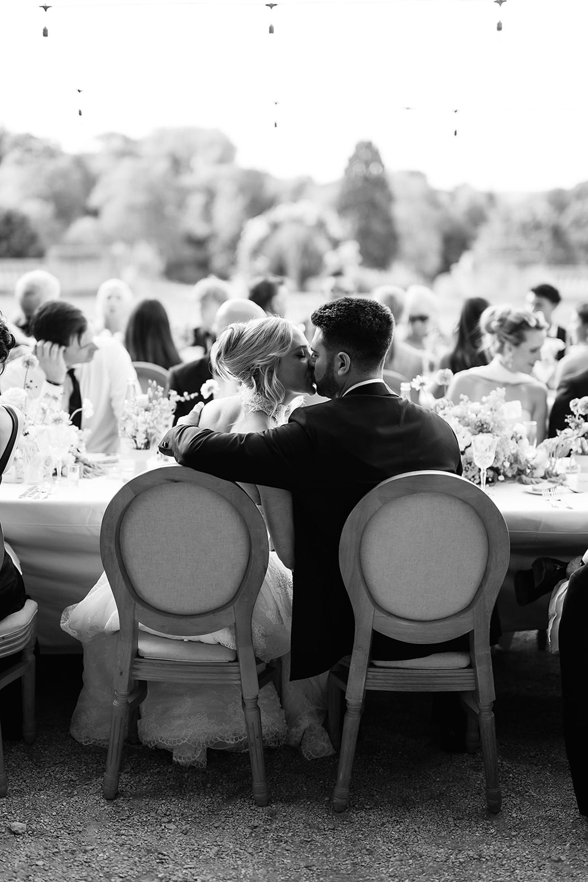 The bride and groom kiss, seen from behind, seated at a long outdoor table with flowers while guests sit beyond them.