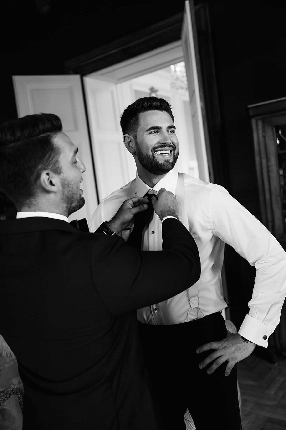 One man in a dark jacket adjusts the bow tie of a smiling man in a white shirt beside a window.