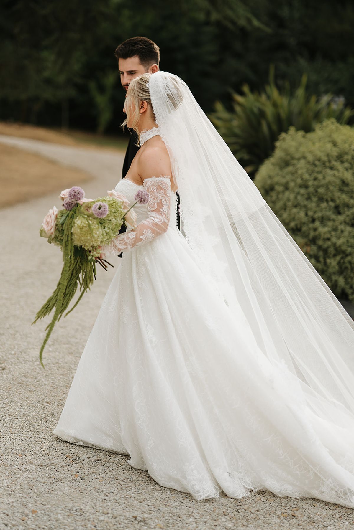The bride in a long veil and lace-sleeved gown holds a bouquet as the groom stands close behind her on a gravel path.
