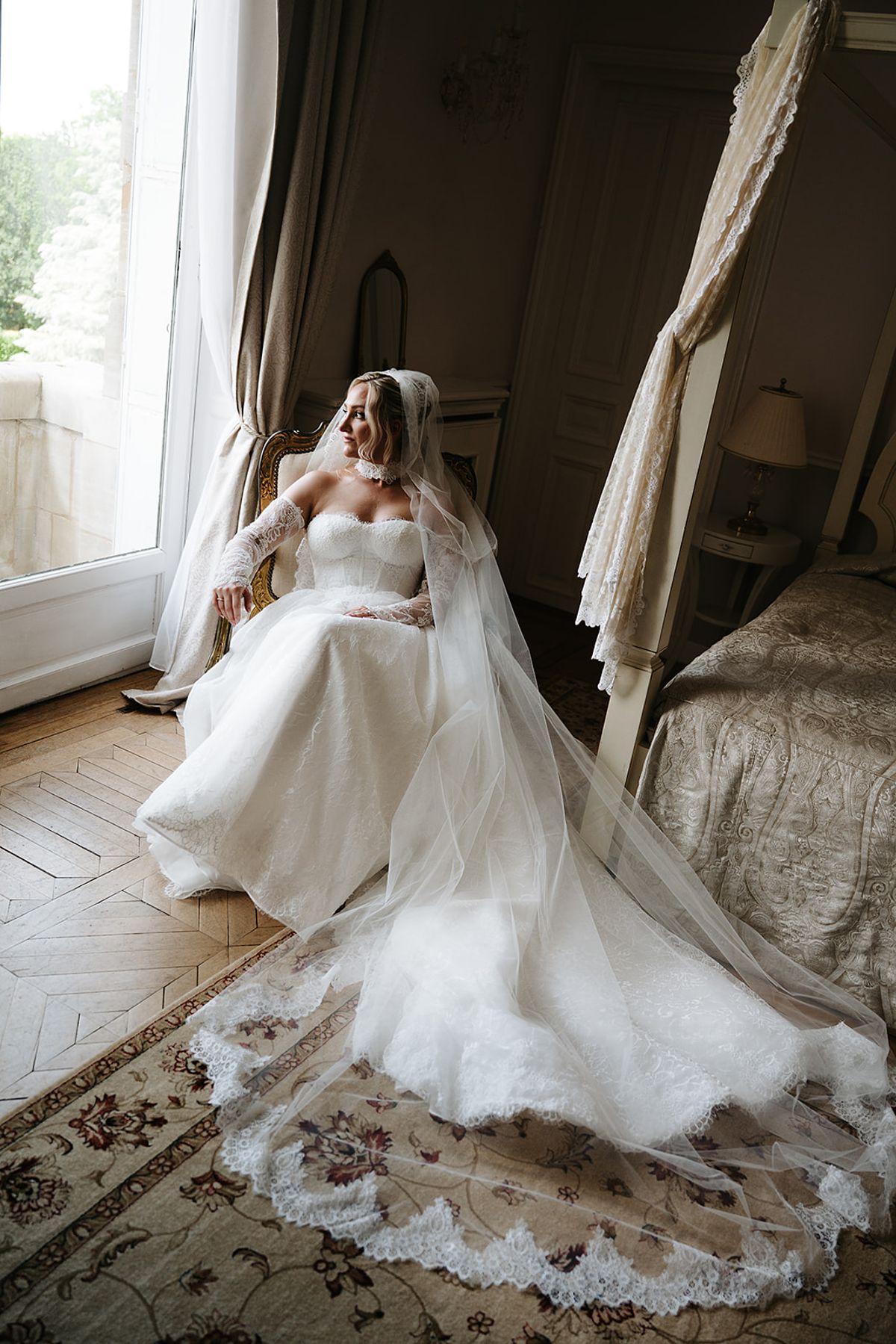 The bride sits in a chair by a tall window in her gown, her long lace-trimmed veil spread across a patterned rug.