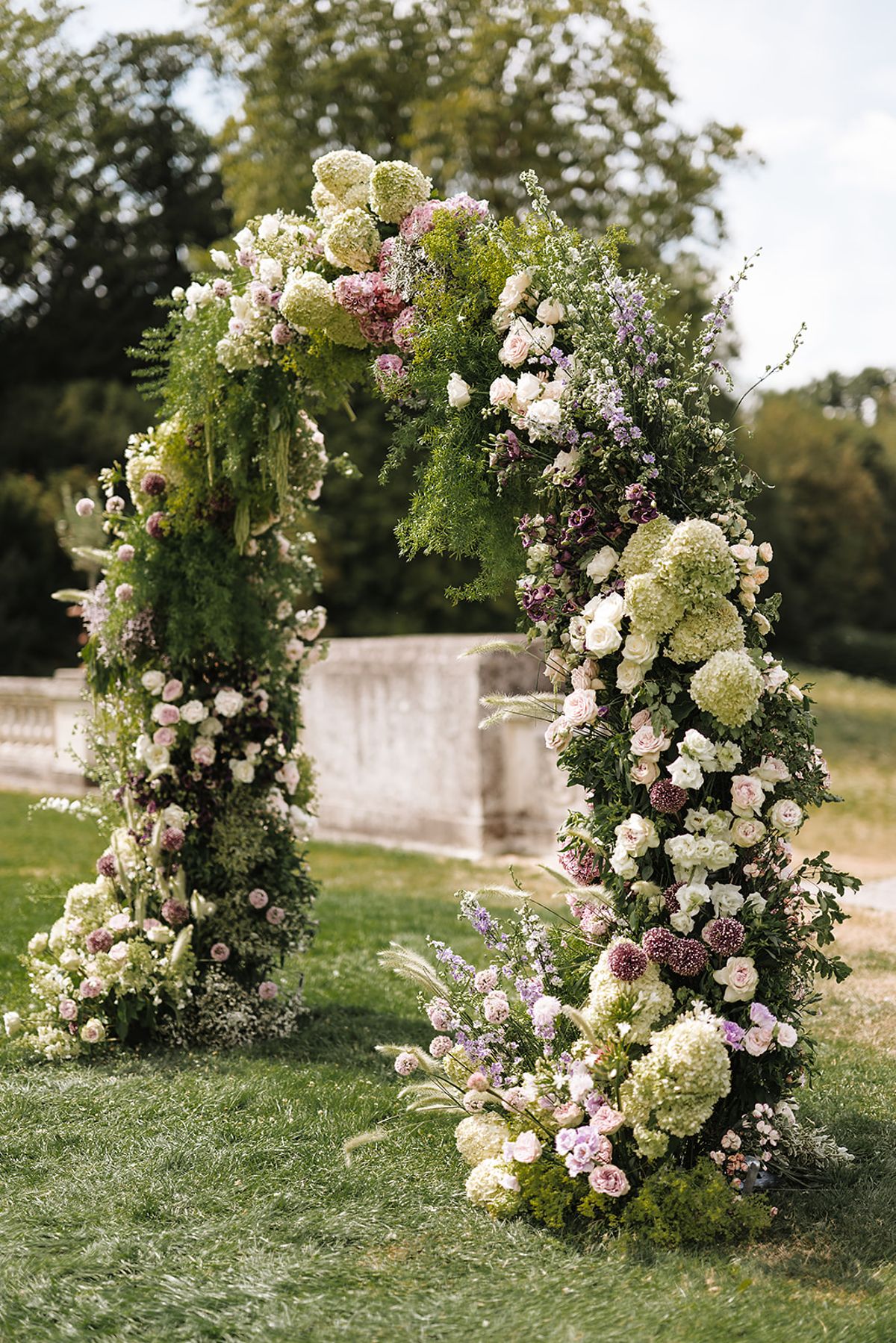 A tall arch covered in white, green and lilac flowers stands on a lawn, with trees and a stone wall behind.