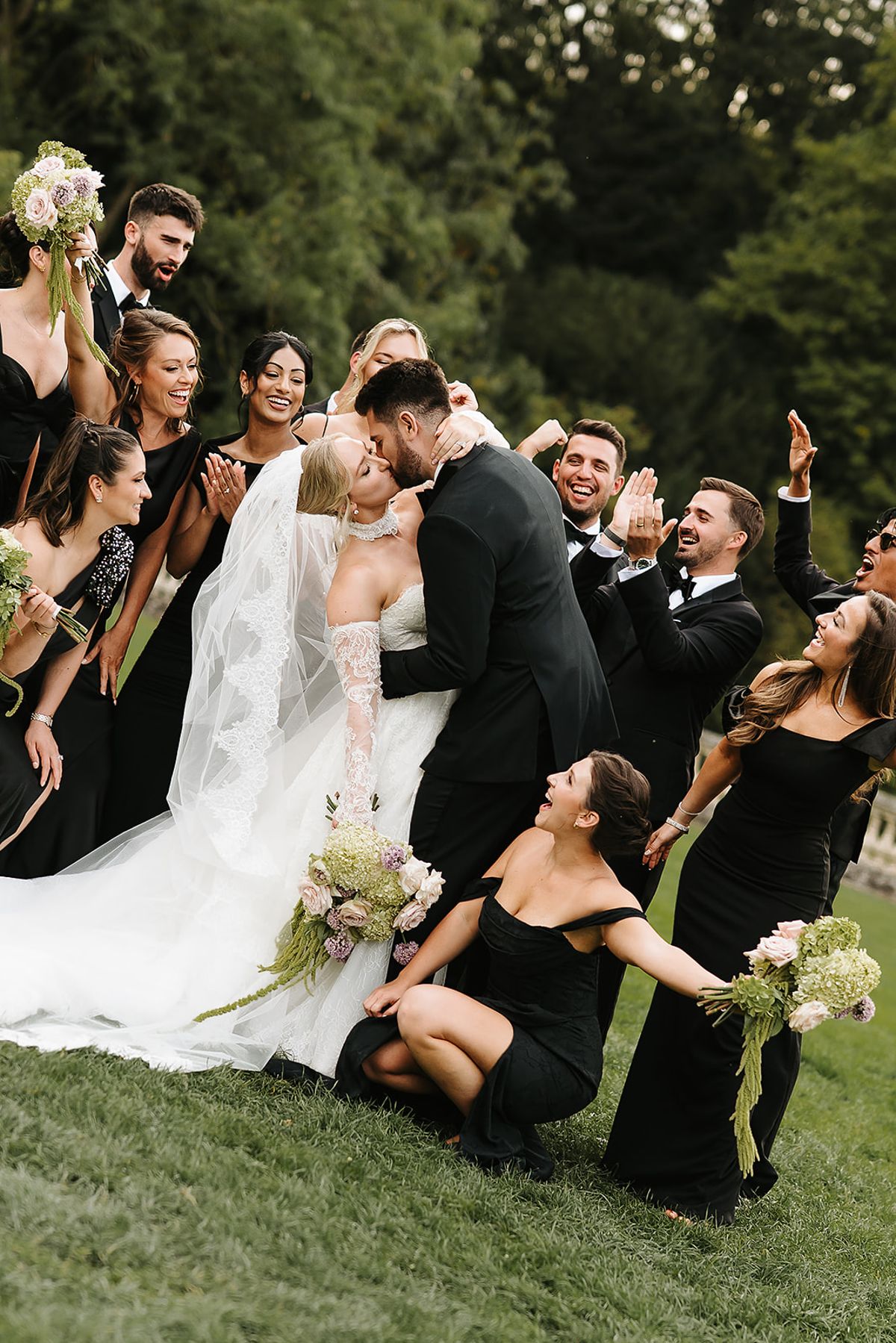 Bridesmaids and groomsmen in black crowd around the bride and groom as they kiss on the grass, laughing and cheering.
