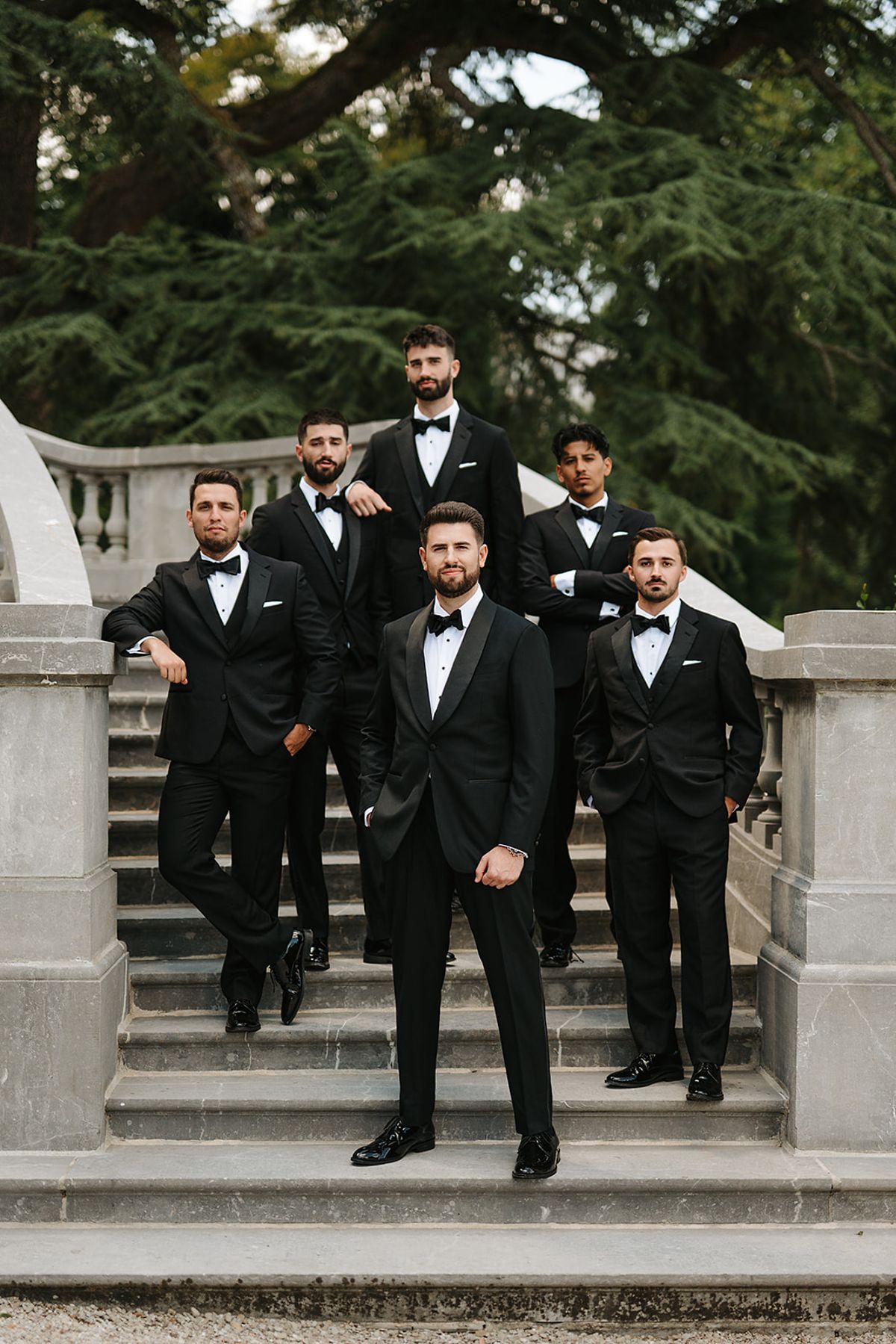 The groom stands at the front of a stone staircase, flanked by six groomsmen in black tuxedos and bow ties.