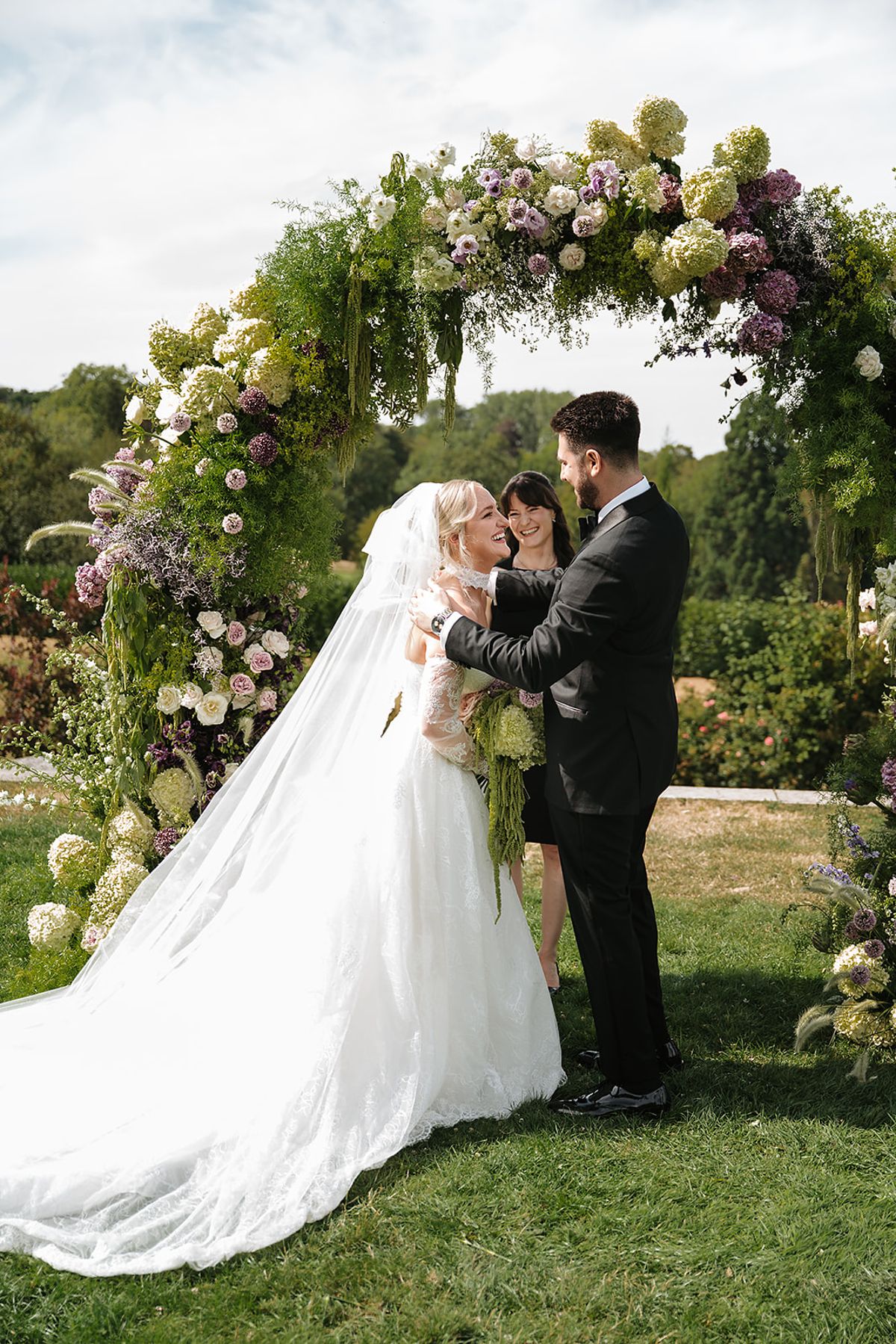 The bride and groom stand facing each other under a round arch of lilac and green flowers on a lawn.