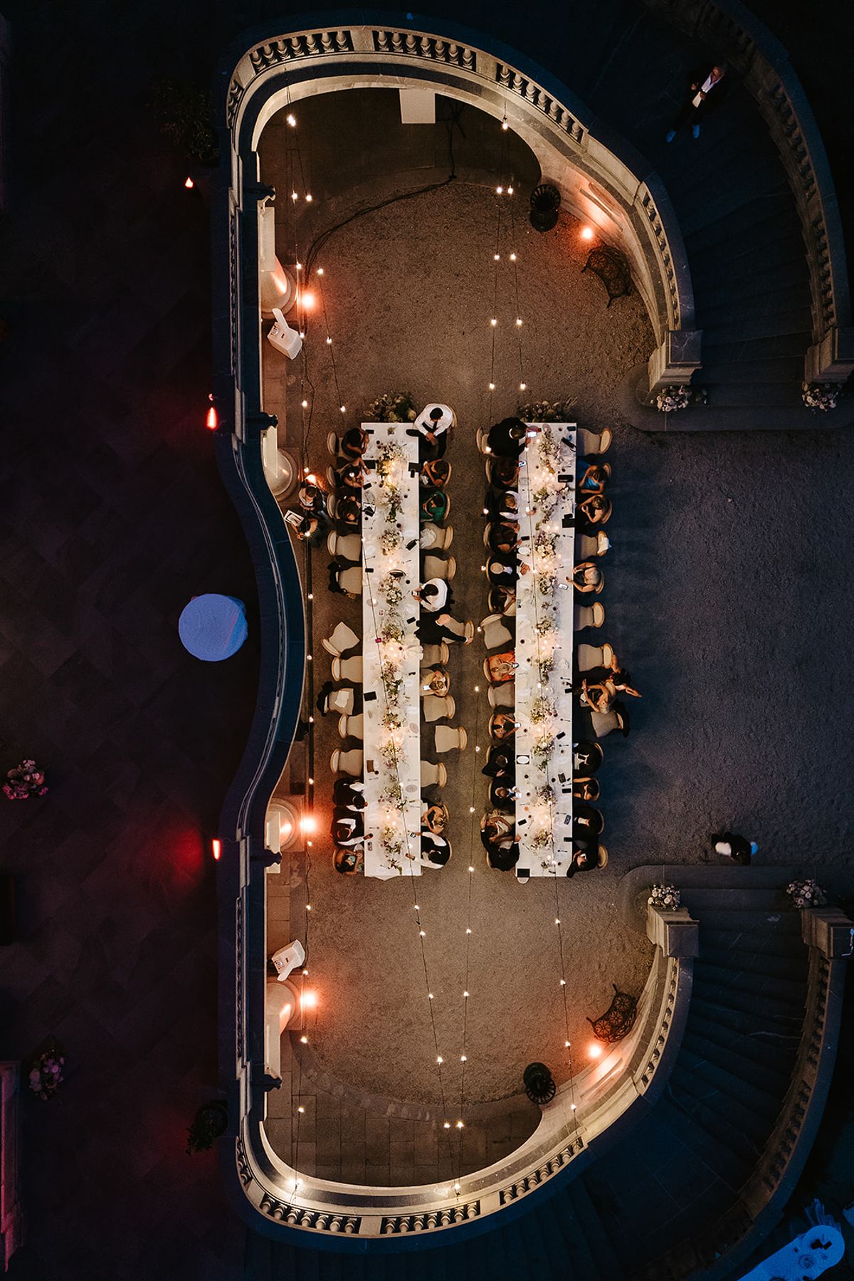 Seen from above at night, guests sit along two long white tables on a curved stone terrace lit by string lights.