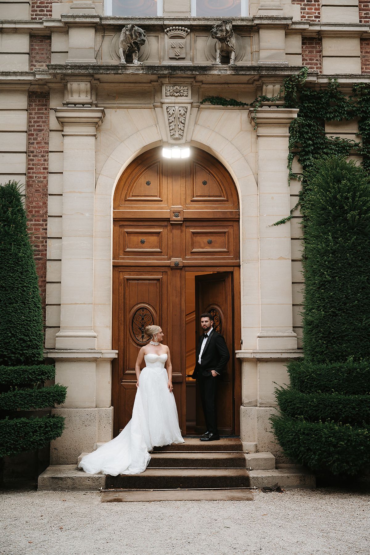 The couple stand on stone steps before a large arched wooden door in a brick and stone chateau wall covered with ivy.