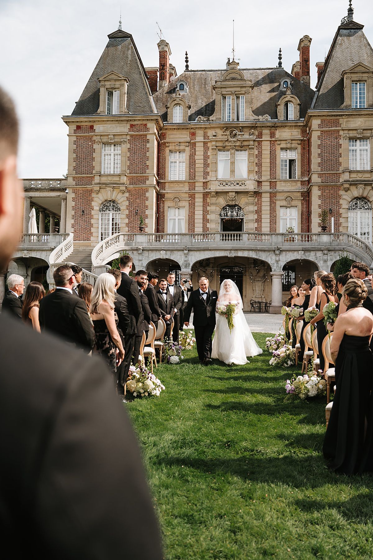 The bride walks down a grass aisle between seated guests, with a large brick chateau in the background.