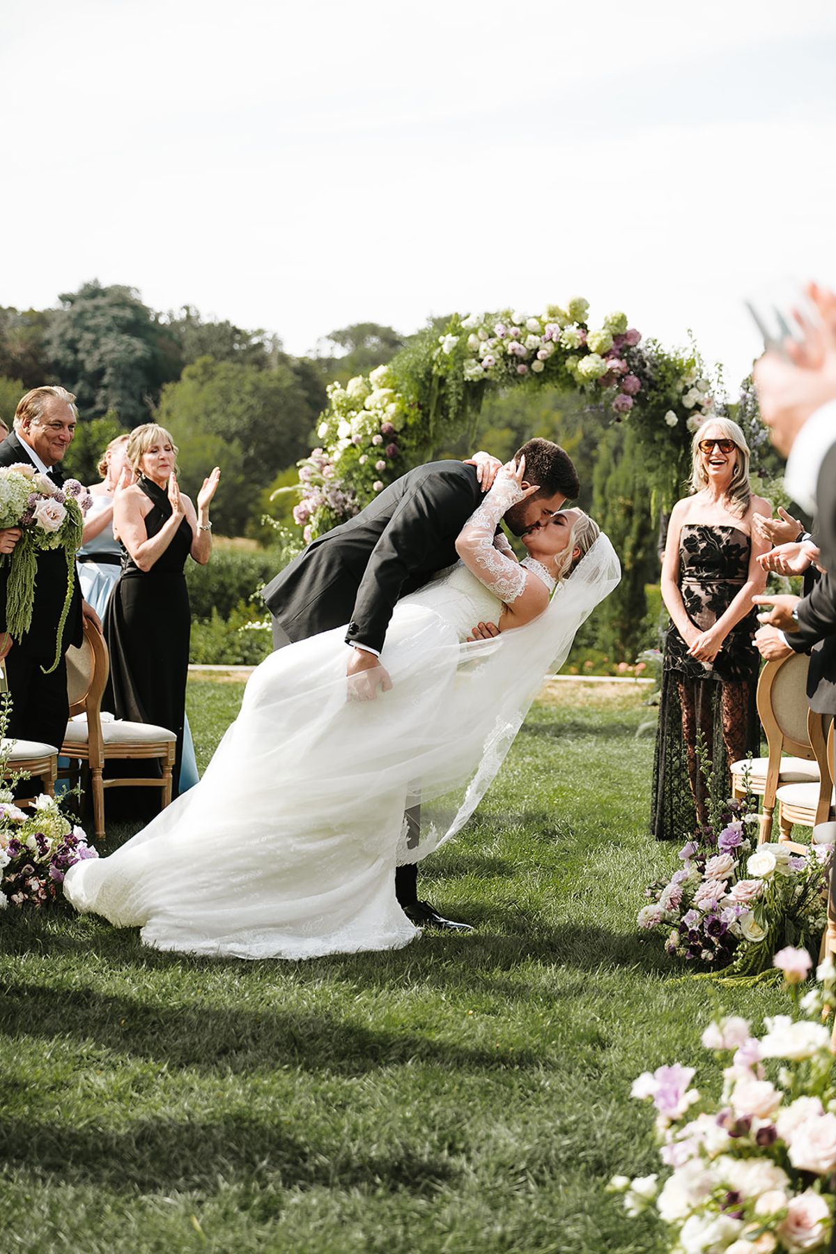 The groom dips the bride for a kiss beneath a floral arch while smiling guests applaud behind them.
