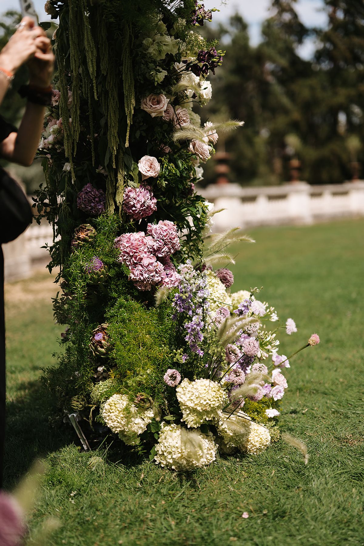 A tall column of pink, lilac and white flowers and greenery stands on a lawn with trees and a stone balustrade behind.