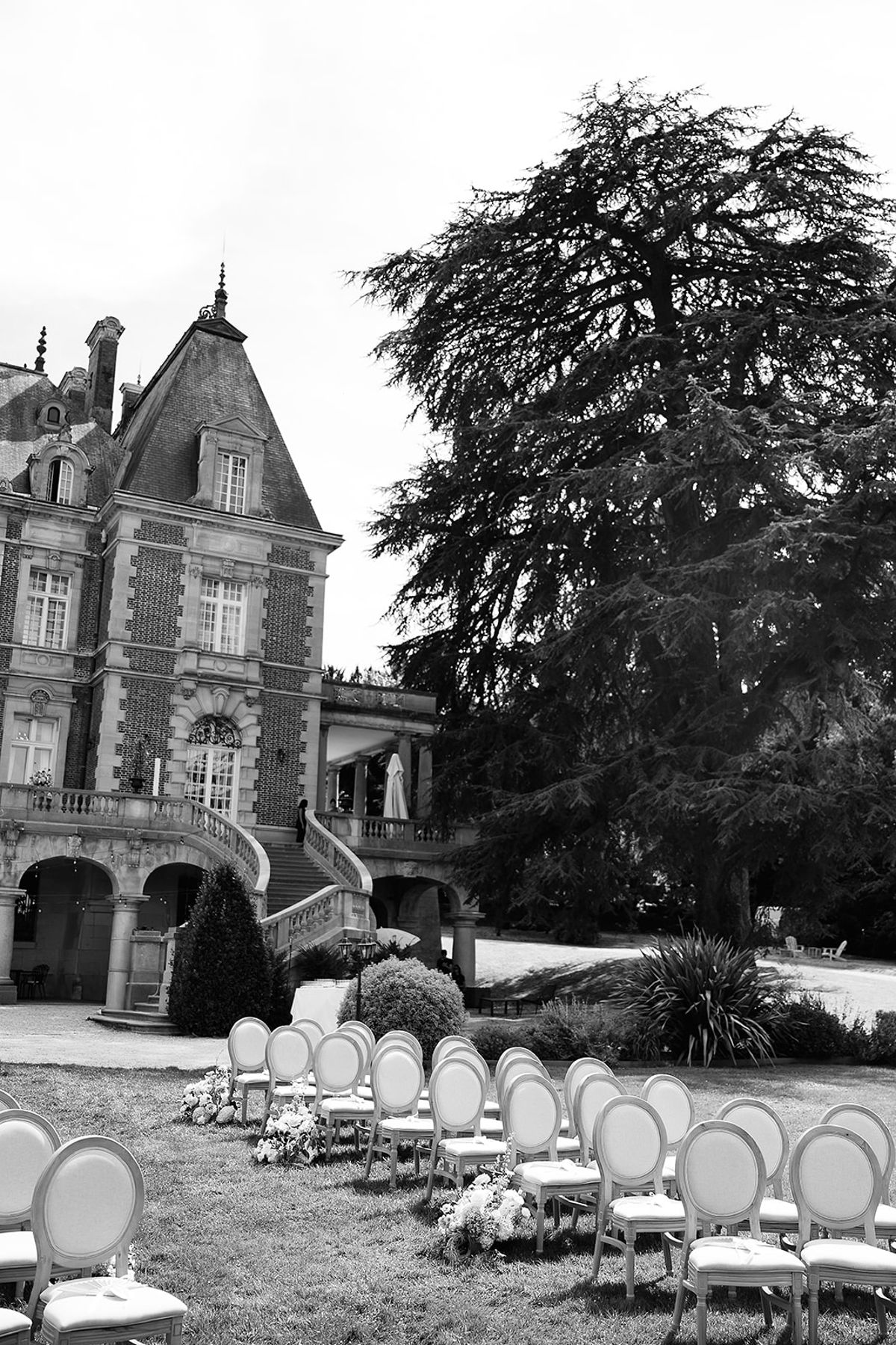 A black and white view of a turreted chateau beside a large tree, with rows of round-backed chairs set out on the lawn.