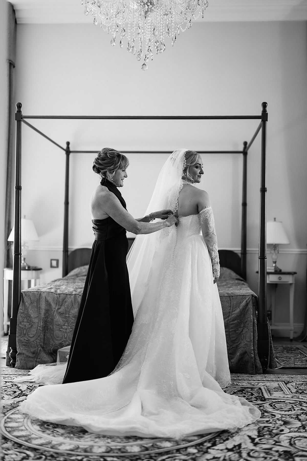A woman in a dark dress fastens the back of the bride's gown in a bedroom with a four-poster bed.