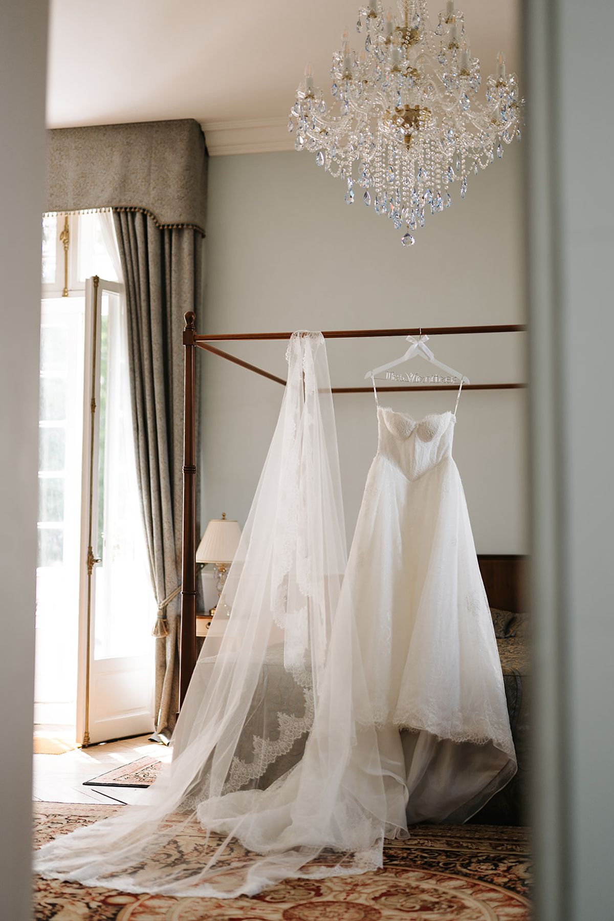 A white wedding dress and long veil hang near a tall window in a bedroom with a crystal chandelier.