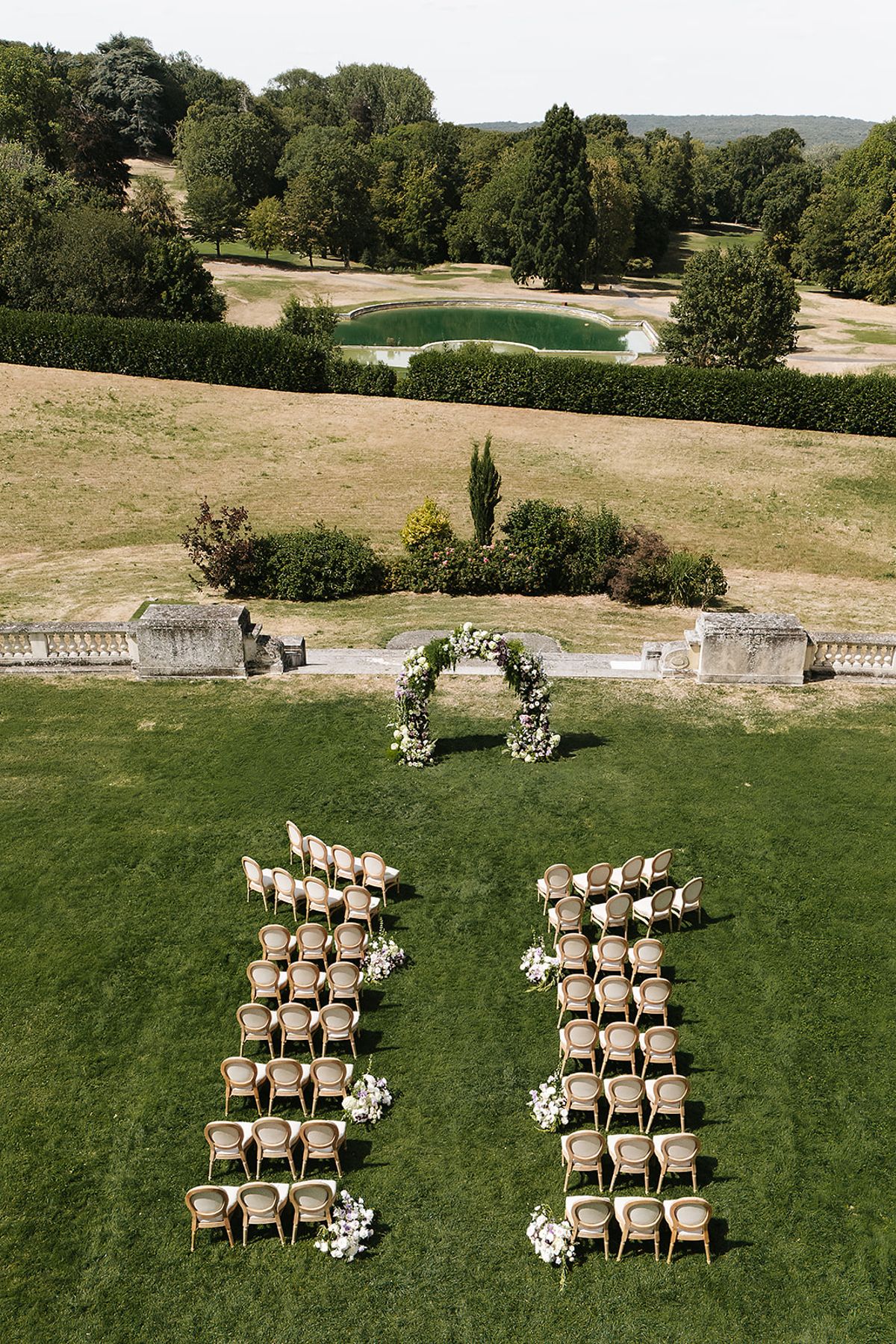 From a raised view, two blocks of wooden chairs face a small floral arch on a green lawn, with a pond and trees beyond.