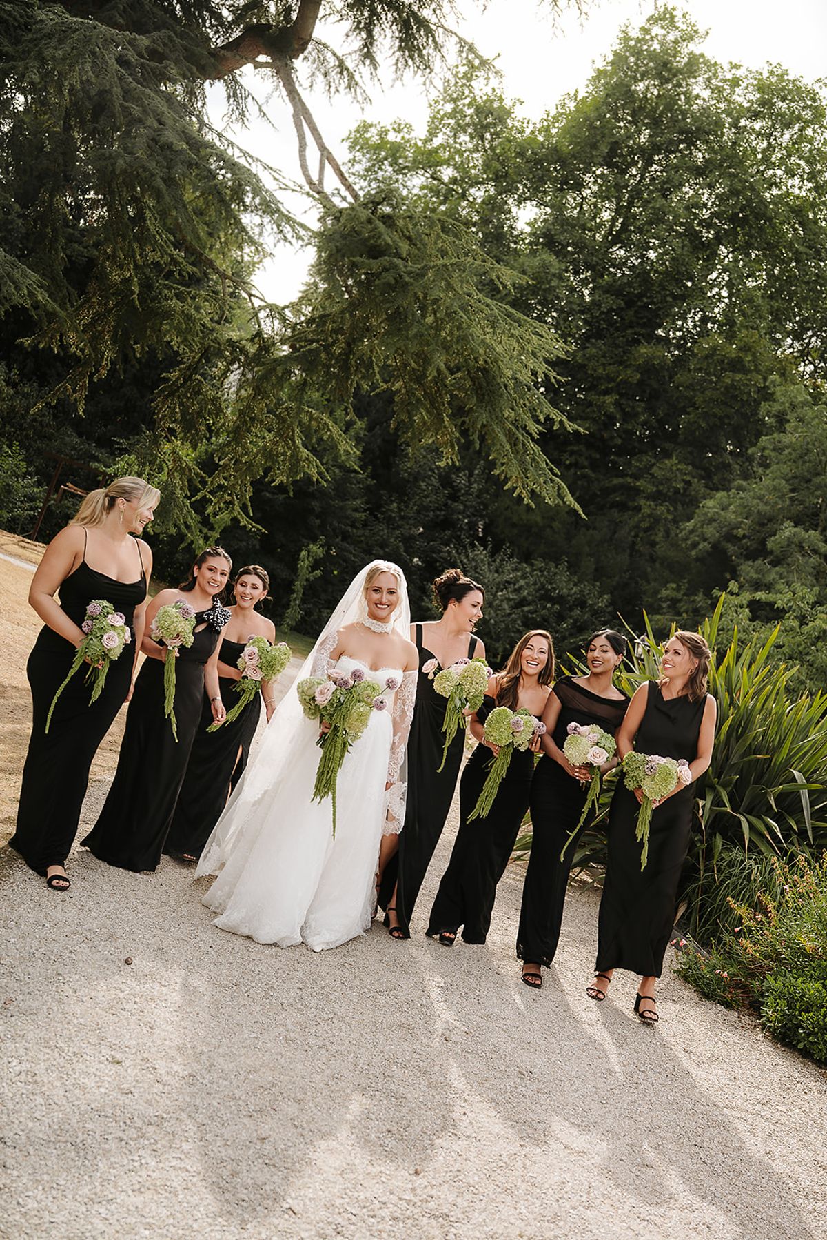 The bride in a white gown walks along a gravel path among bridesmaids in long black dresses, each carrying green and white flowers.