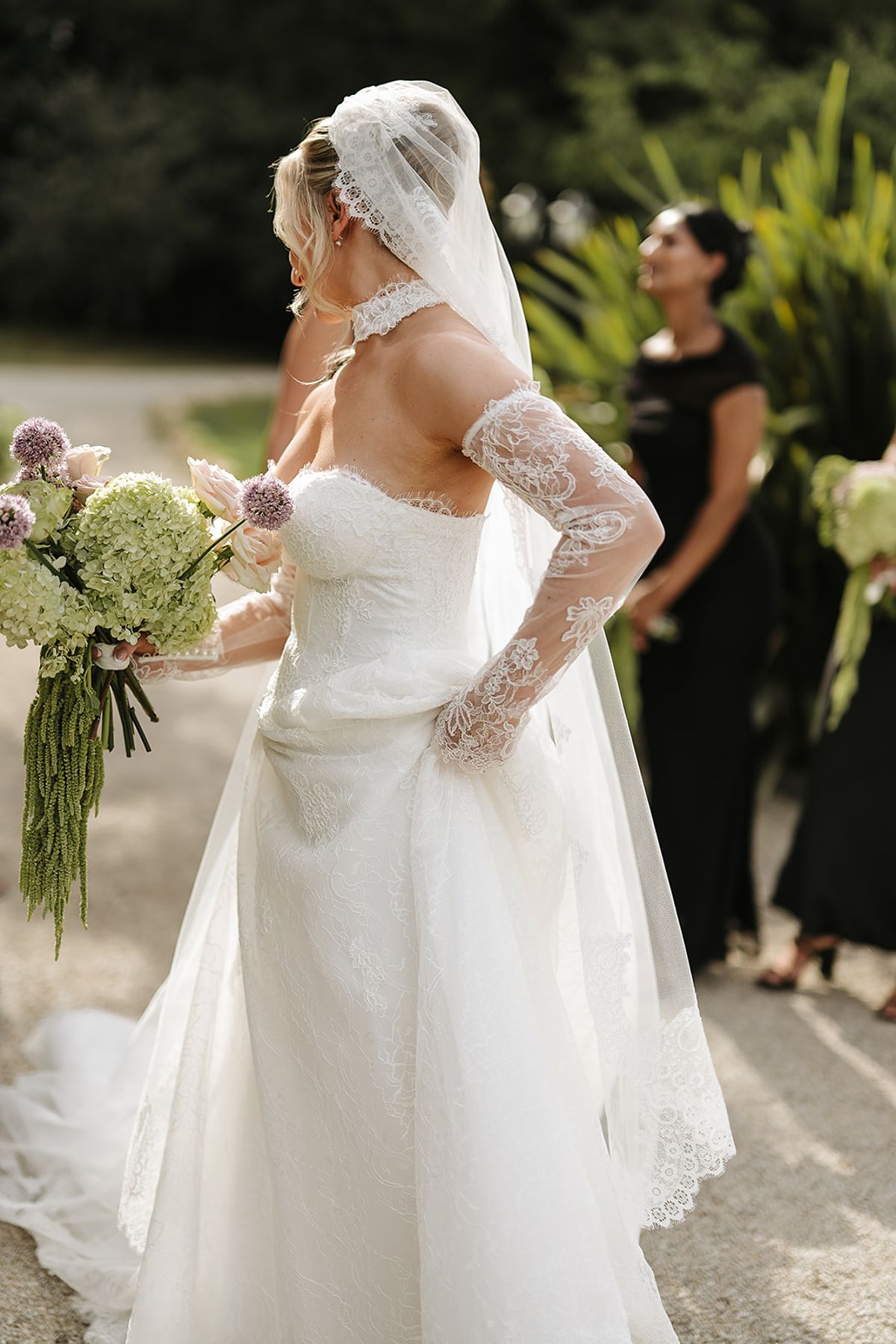 The bride in a strapless lace gown, long lace gloves and veil holds a green and lilac bouquet; guests in black stand behind.