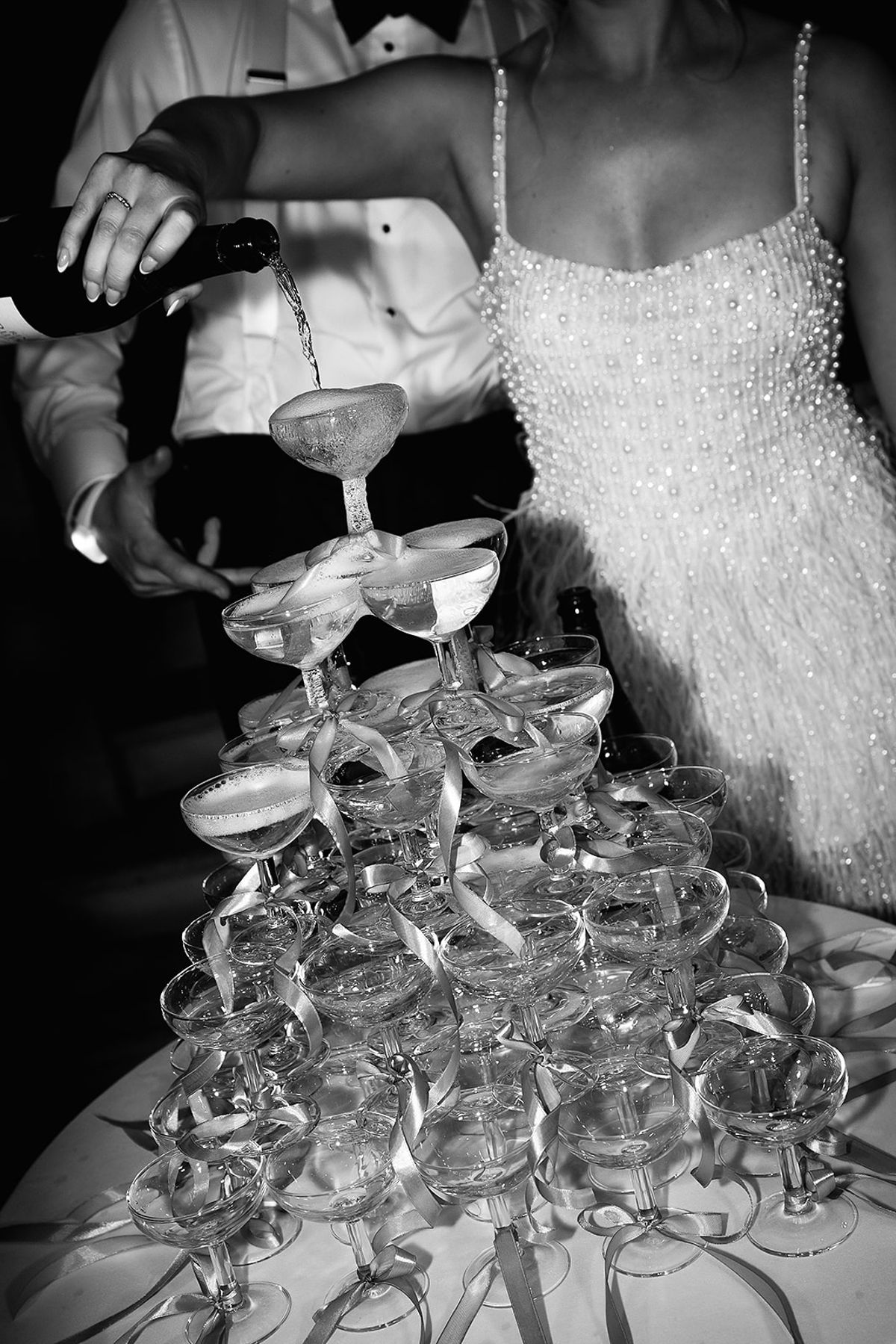 In black and white, champagne is poured into the top coupe of a ribbon-tied glass tower, beside a woman in a short pearl-beaded dress with feather trim.