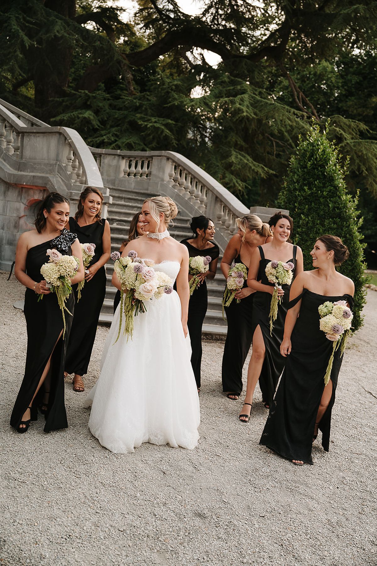 The bride in a white gown walks with seven bridesmaids in long black dresses, carrying pale bouquets, beside a stone staircase.
